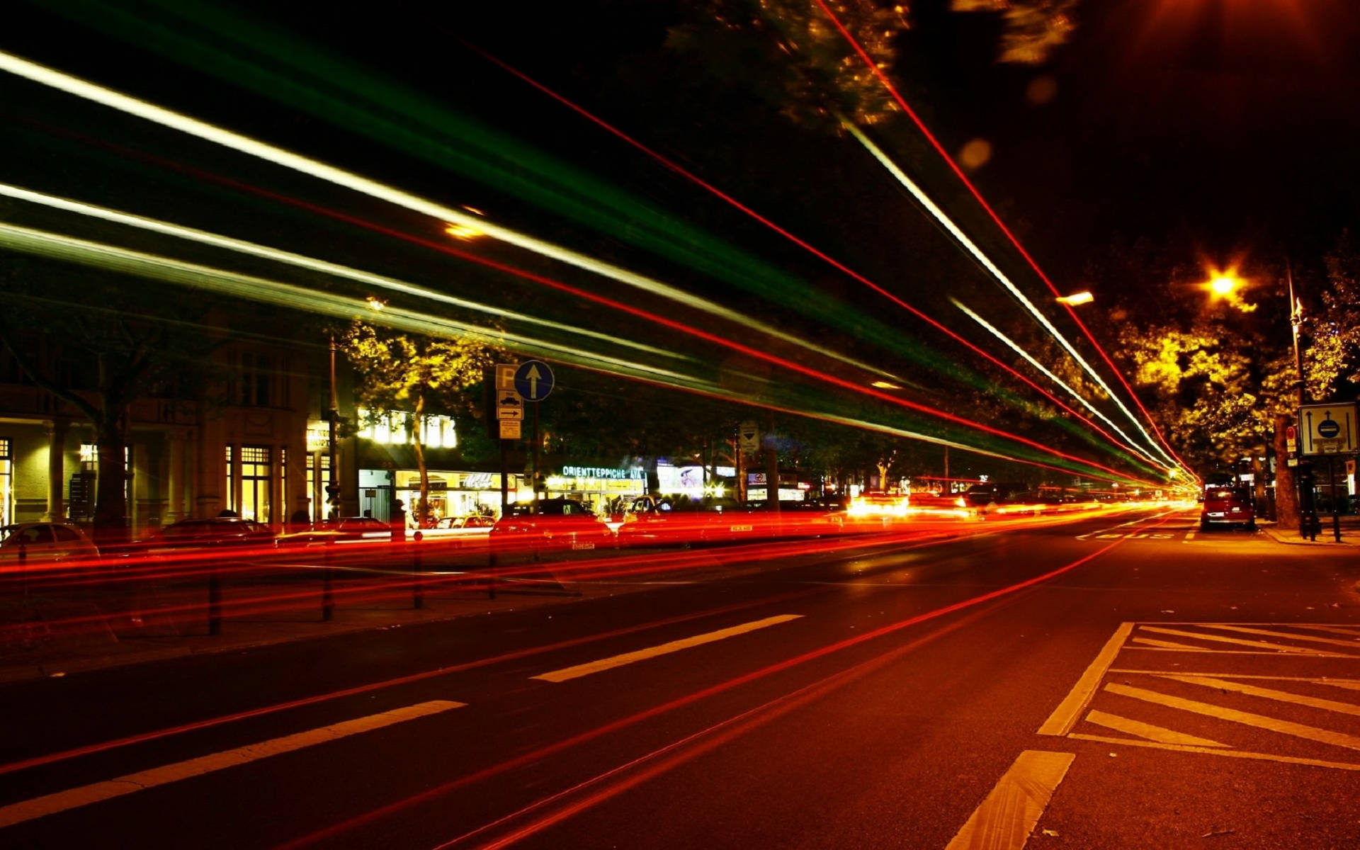 Berlin 桌布 壁紙 — 紅色、綠色和白色的鮮豔光帶在夜間濕漉漉的城市街道上模糊地掠過，捕捉到發光的商店櫥窗和樹木前的動感。