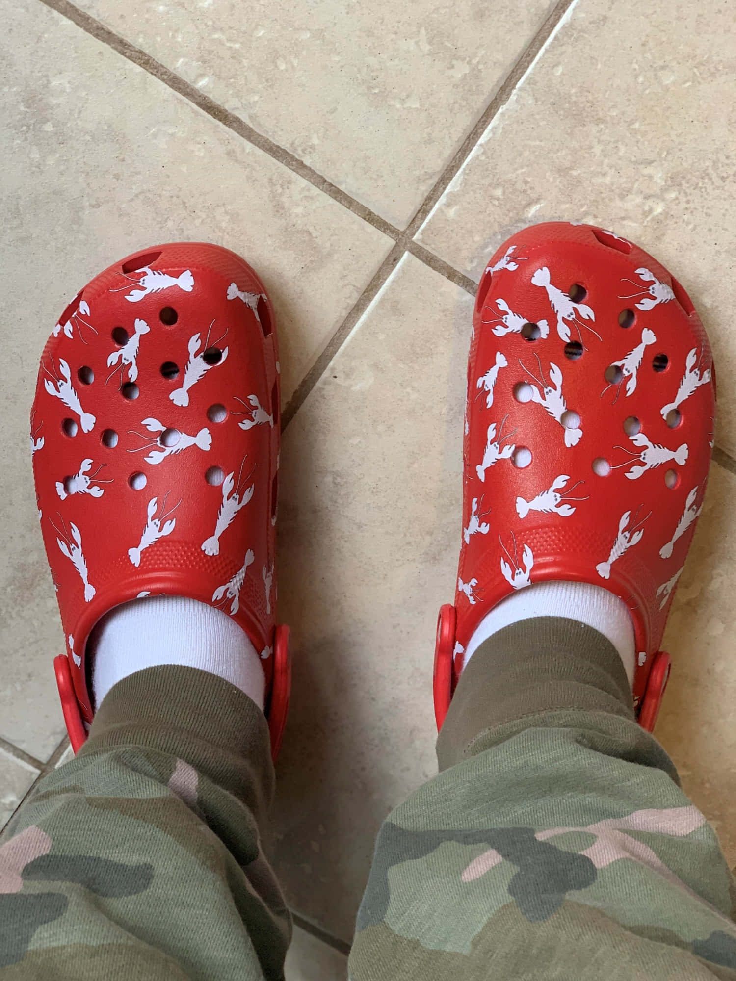Crocs 桌布 壁紙 — 紅色Crocs鞋帶有白色龍蝦圖案，放在米色瓷磚地板上，搭配迷彩褲和白色襪子。