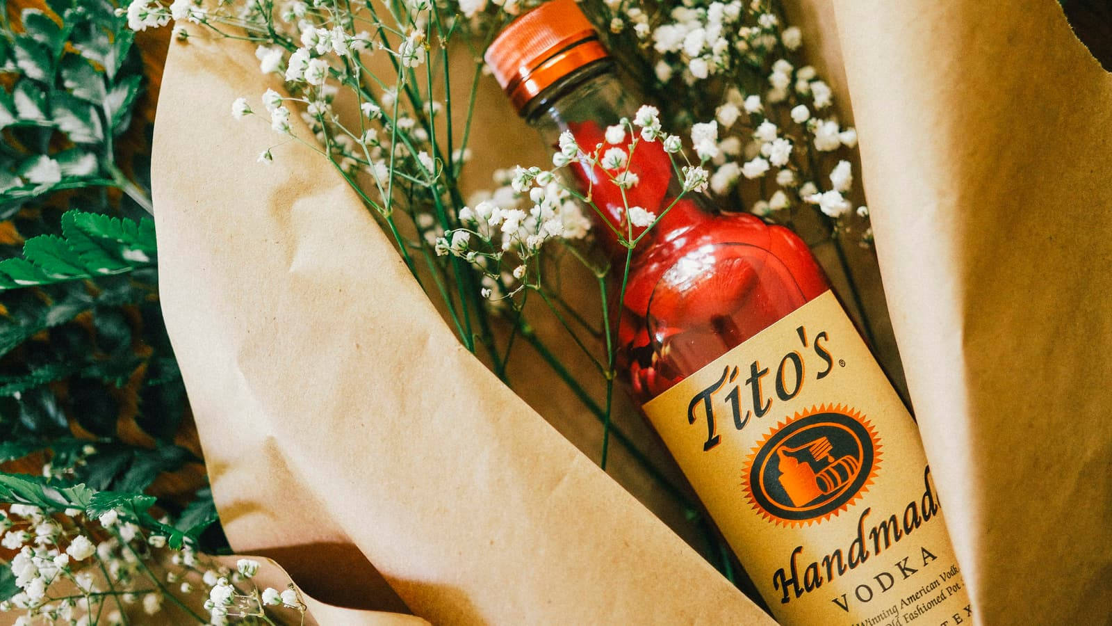 alcohol 桌布 壁紙 — 紅色Tito's伏特加酒瓶藏在牛皮紙和滿天星花及綠色蕨類植物中。