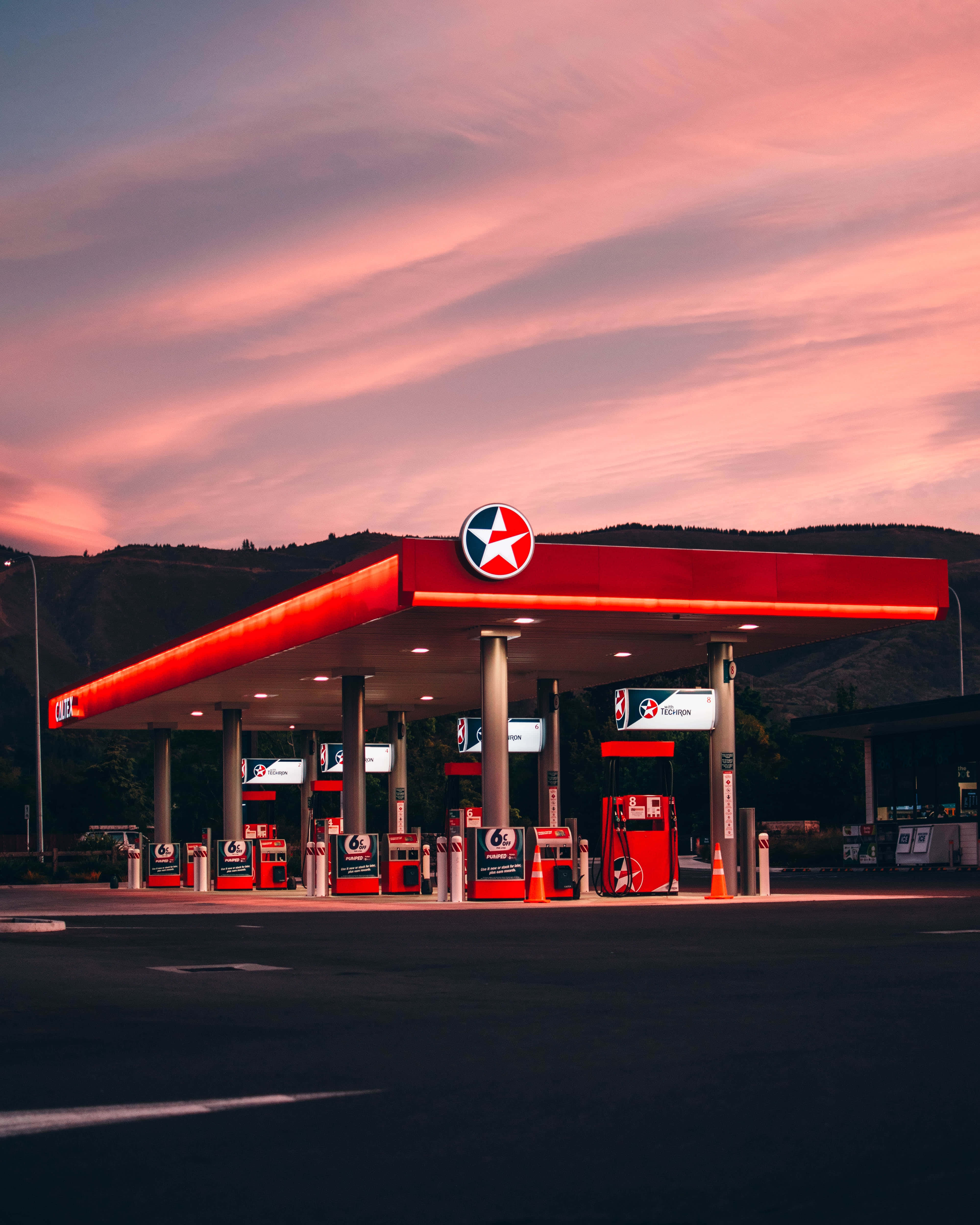 Gas Station 桌布 壁紙 — 紅色加油站頂篷在粉紅和橙色日落天空下發光，背景有遠山。