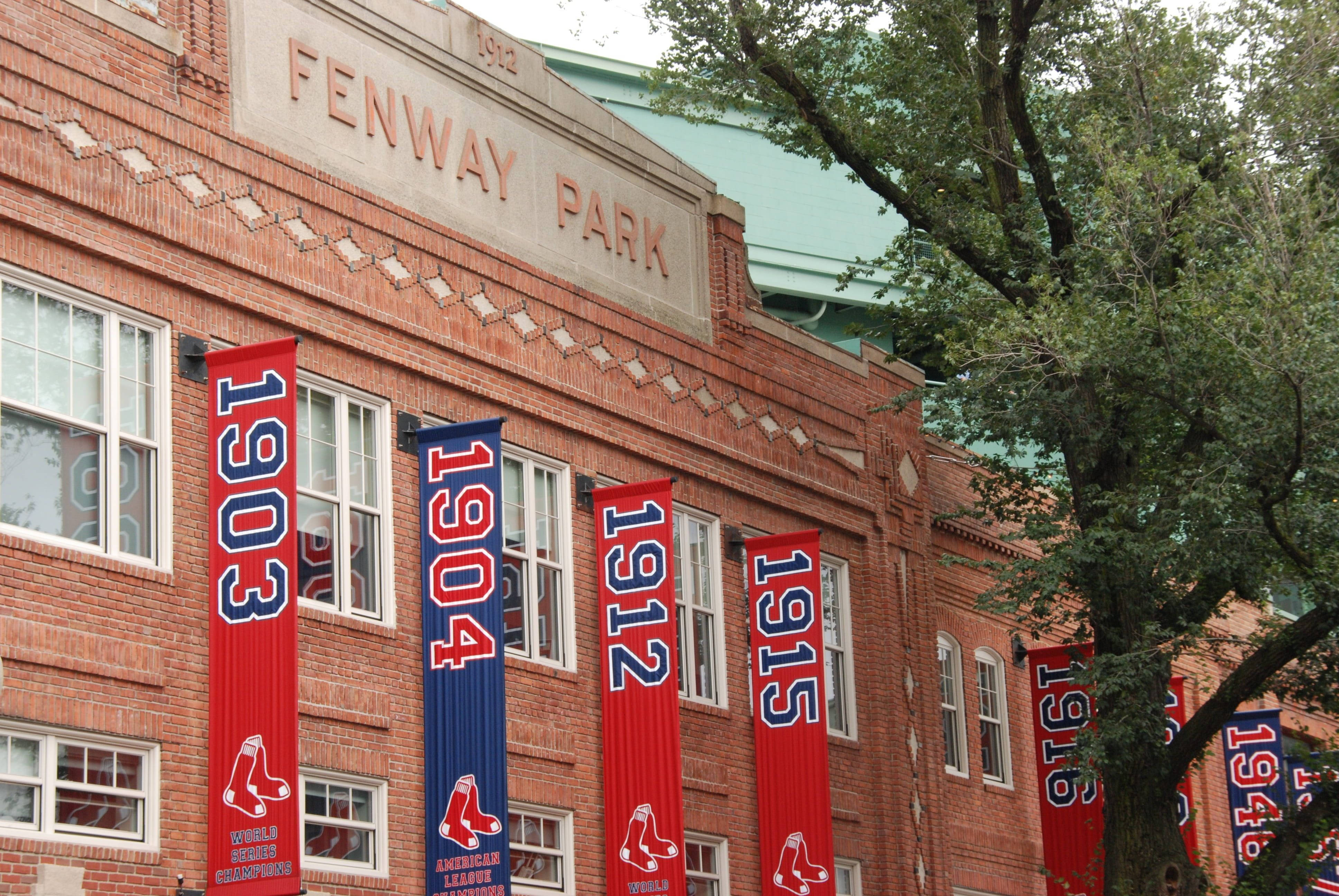 Boston Red Sox 桌布 壁紙 — 紅色和藍色的冠軍旗幟懸掛在芬威球場歷史悠久的磚砌外牆上，下方是綠樹成蔭。