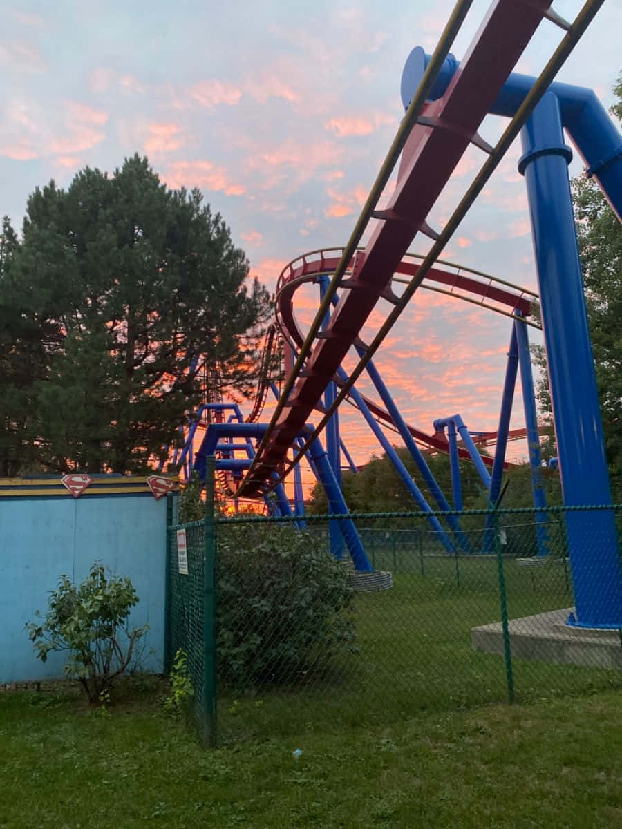 Six Flags 桌布 壁紙 — 紅色和藍色的雲霄飛車在樹木和鐵絲網圍欄映襯下的鮮豔日落天空中蜿蜒。