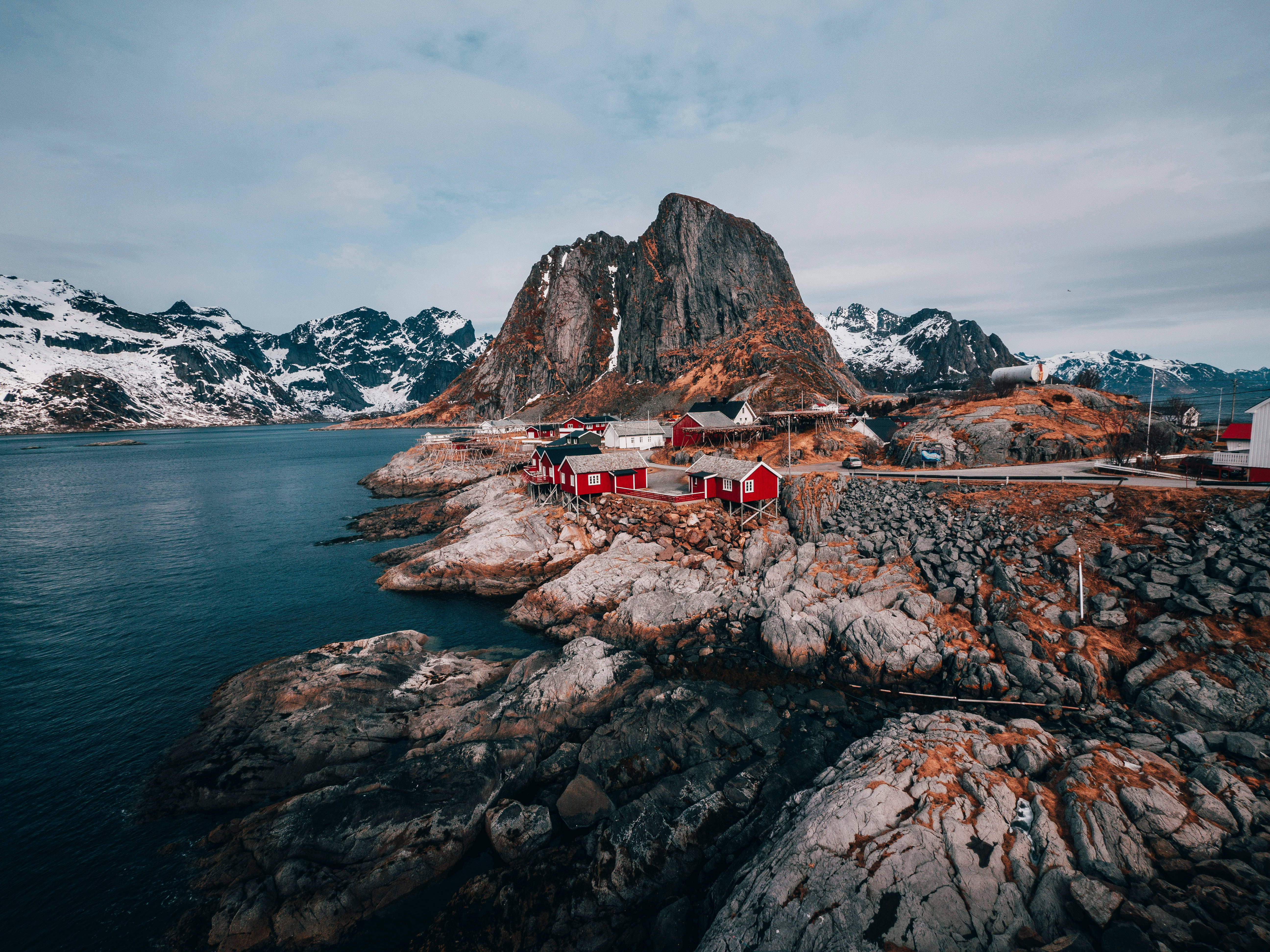 Norway 桌布 壁紙 — 紅色小屋依附在岩石海岸旁，深藍色的水邊，背後有積雪覆蓋的山脈在陰天升起。