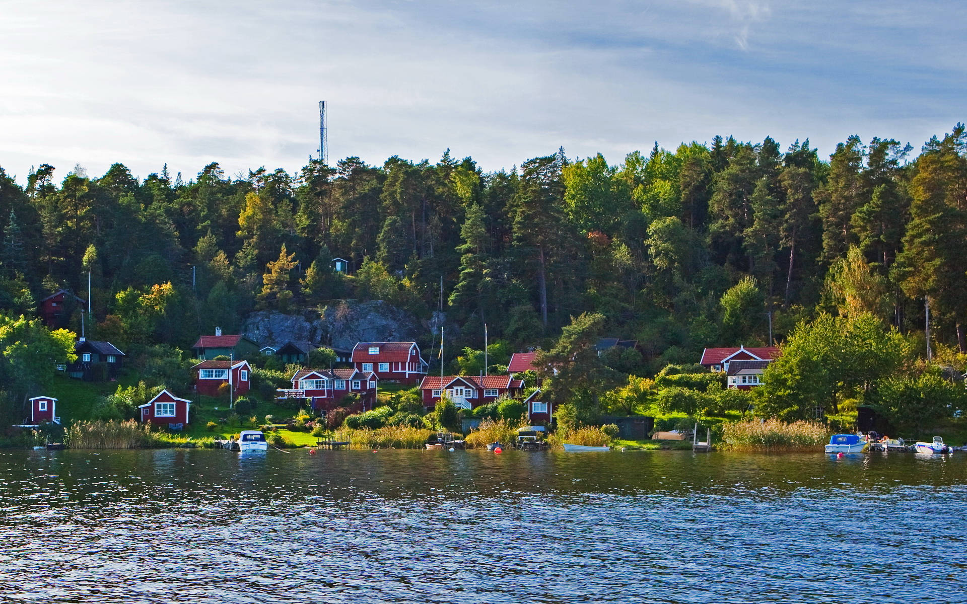 Sweden 桌布 壁紙 — 紅色木屋點綴著樹木繁茂的湖岸，天空部分多雲，船隻在平靜的水面上休息。