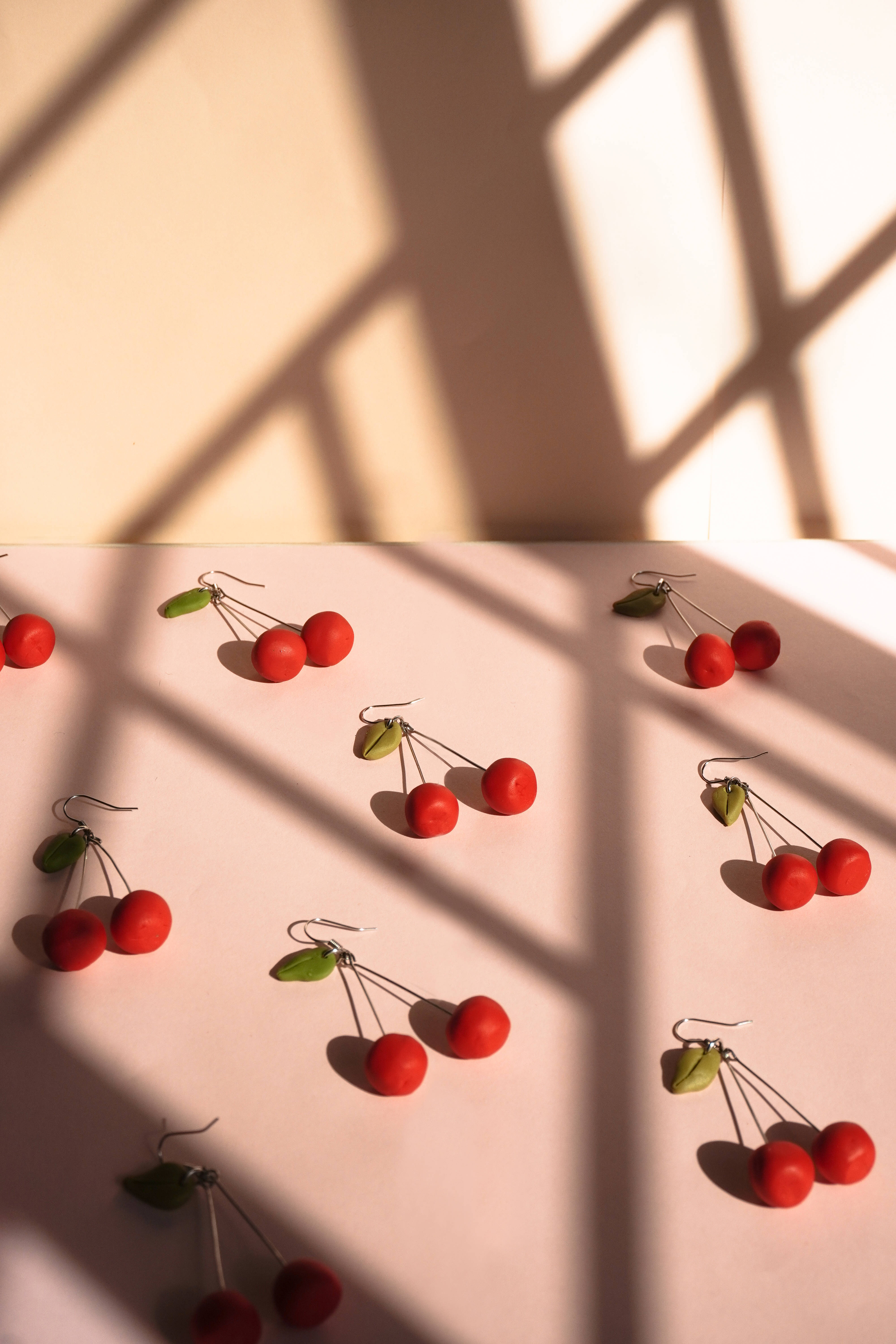 Cute Cherry Aesthetic 桌布 壁紙 — 紅色櫻桃耳環在溫暖的窗光下，於淡粉色表面上投下柔和陰影。