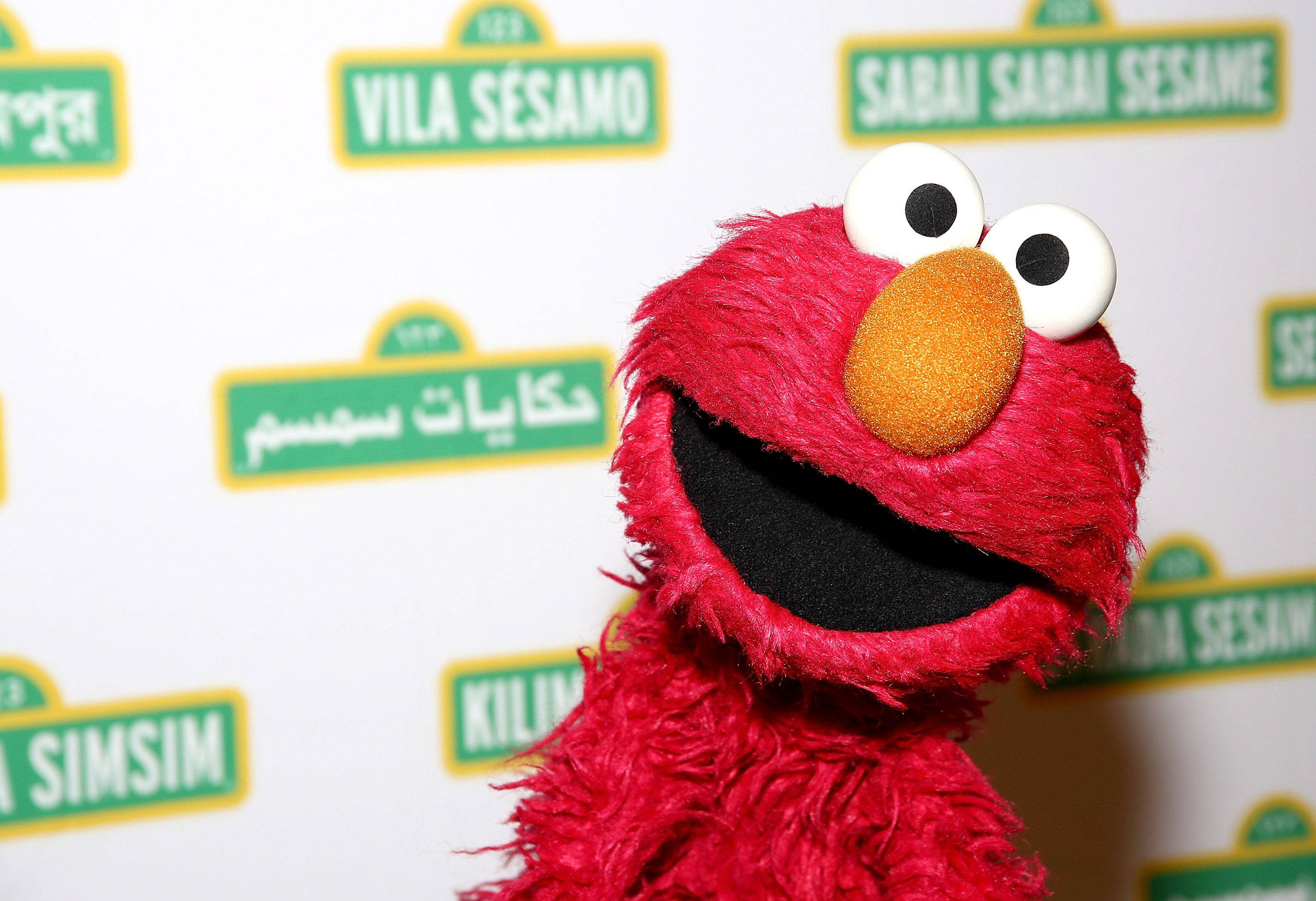 Elmo 桌布 壁紙 — 紅色毛絨絨的 Elmo 木偶在白色背景前咧嘴大笑，背景有多語言的芝麻街標誌。