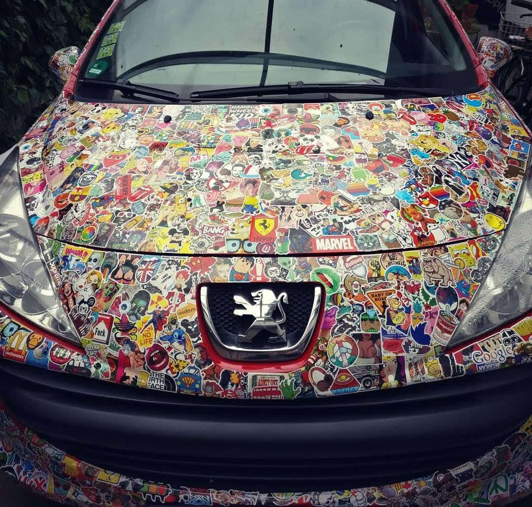 動漫車 桌布 壁紙 — 紅色汽車的引擎蓋上密密麻麻地貼滿了色彩繽紛的卡通和流行文化貼紙，包括Marvel和Peugeot標誌。