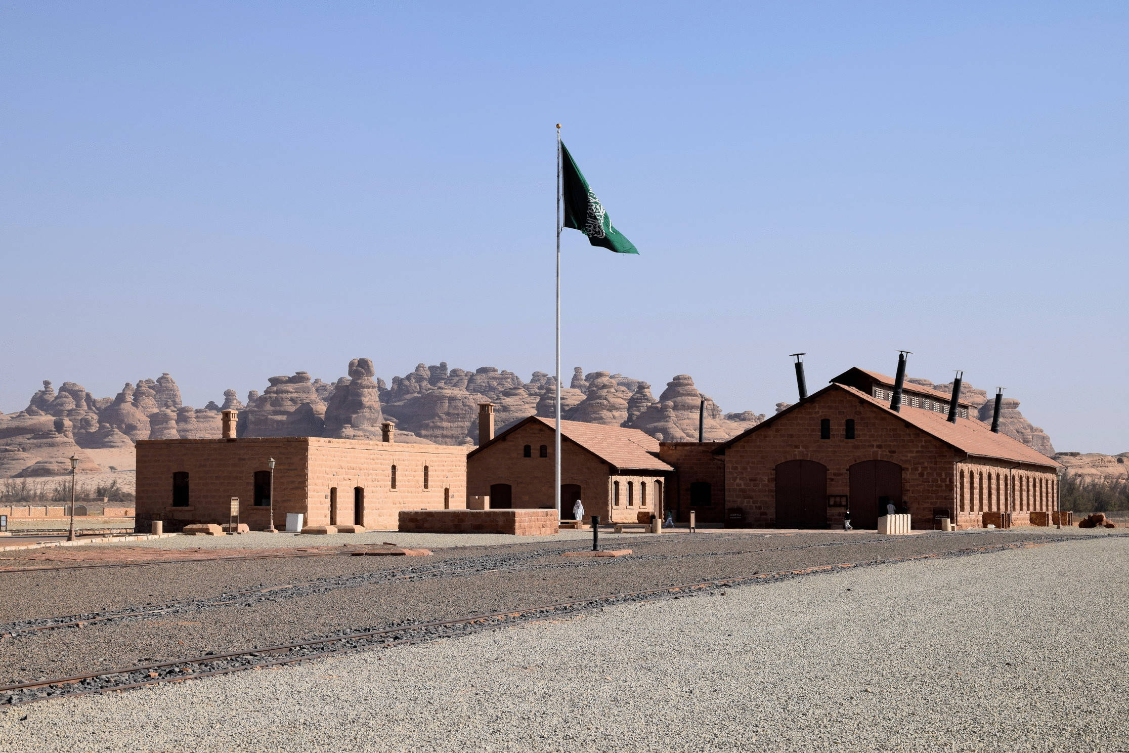 Saudi 桌布 壁紙 — 紅色磚建築矗立在沙漠中，一面綠色旗幟在清澈的藍天和岩石地貌背景下飄揚。