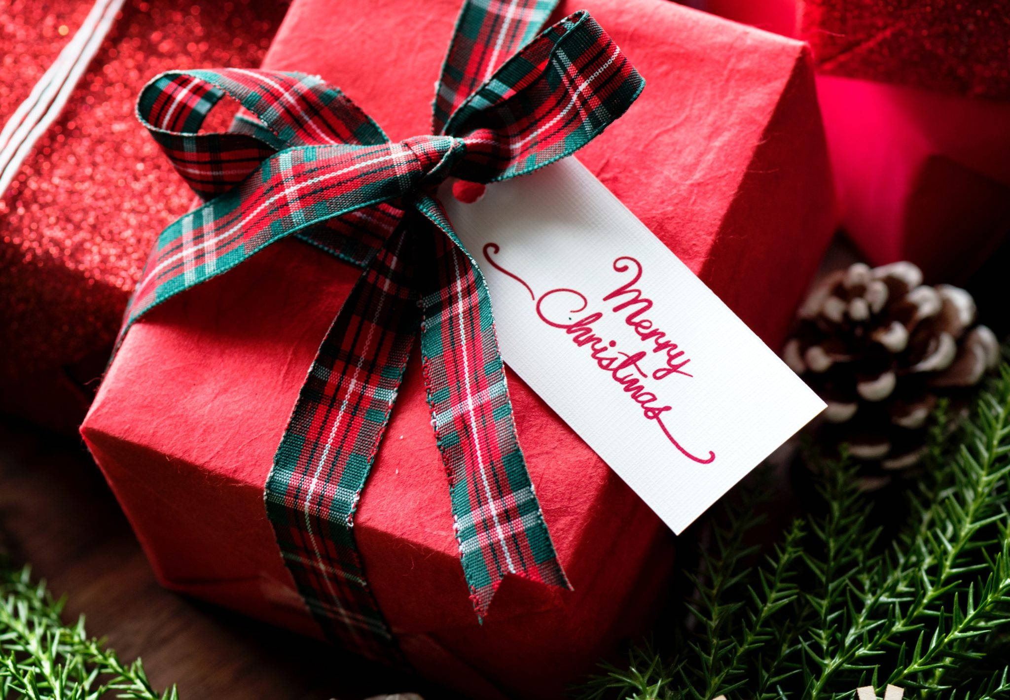 Presents 桌布 壁紙 — 紅色禮物盒的特寫，繫著格子絲帶和「Merry Christmas」標籤，周圍環繞著松果和常青樹枝。