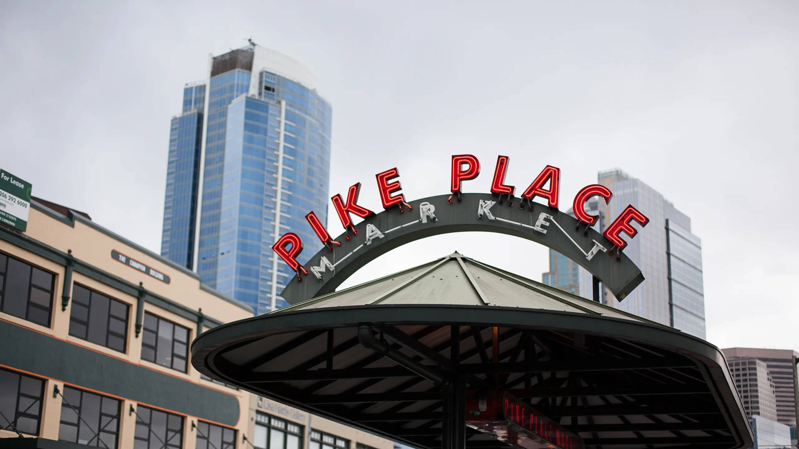 Pike Place Market 桌布 壁紙 — 紅色霓虹燈的Pike Place Market標誌拱在一個有蓋的亭子上方，玻璃摩天大樓在陰天背景下聳立其後。