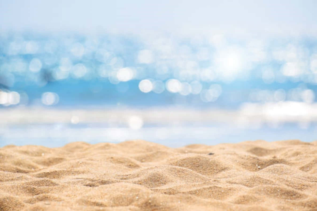 sand 桌布 壁紙 — 紋理金色沙灘的特寫，模糊的藍色海浪在明亮的陽光下閃閃發光。