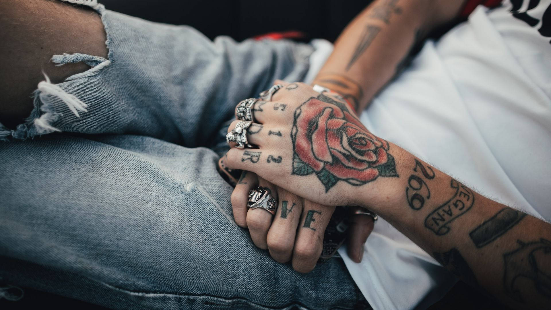 Tattoo 桌布 壁紙 — 紋身雙手緊握，帶有紅玫瑰紋身和戒指，背景是破洞牛仔褲的特寫。