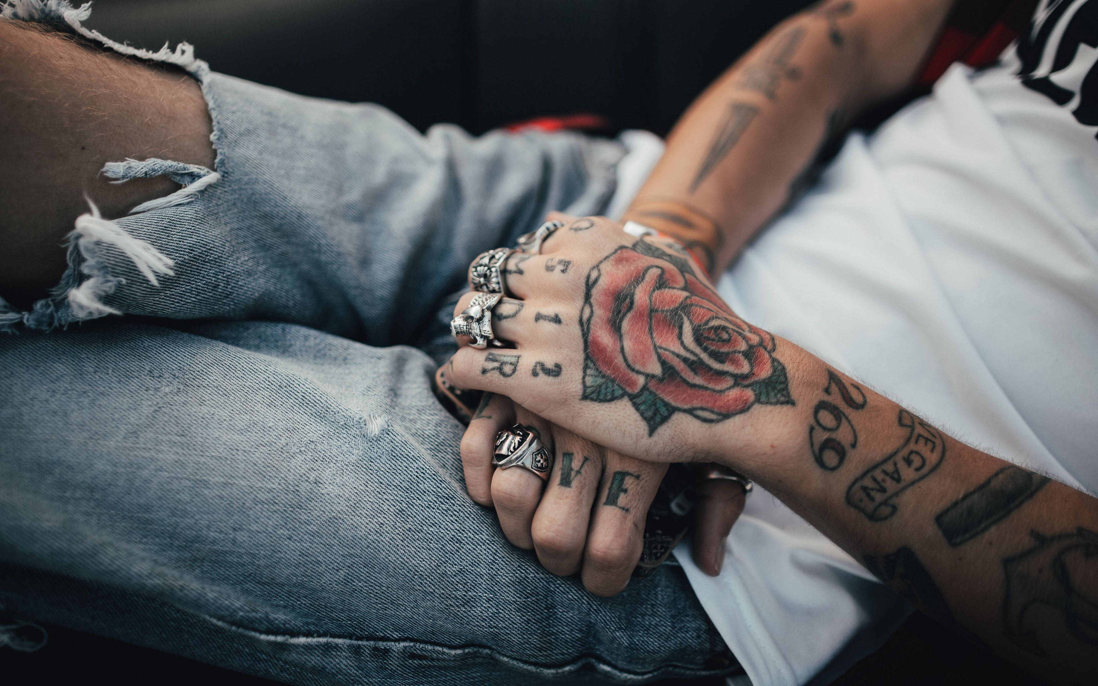 Tattoo 桌布 壁紙 — 紋身雙手緊握，帶有紅玫瑰設計和戒指，背景是破洞牛仔褲的特寫。