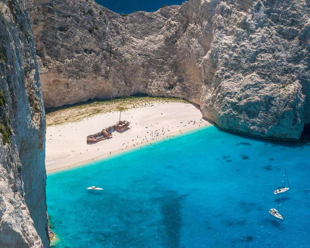 Mediterranean Beach 桌布 壁紙 — 納瓦吉奧海灘的鳥瞰圖，擁有碧綠的海水、白色沙灘、高聳的懸崖和一個生鏽的沉船，周圍有遊客。