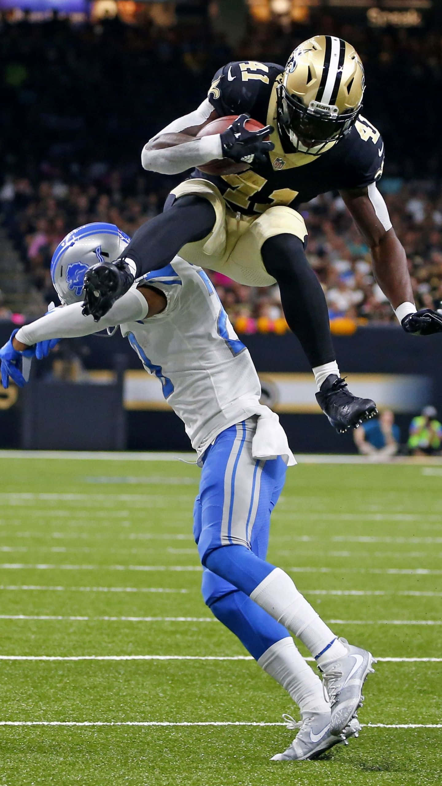 Alvin Kamara 桌布 壁紙 — 紐奧良聖徒隊球員在夜間比賽中跳過底特律雄獅隊防守球員，同時緊抱橄欖球。