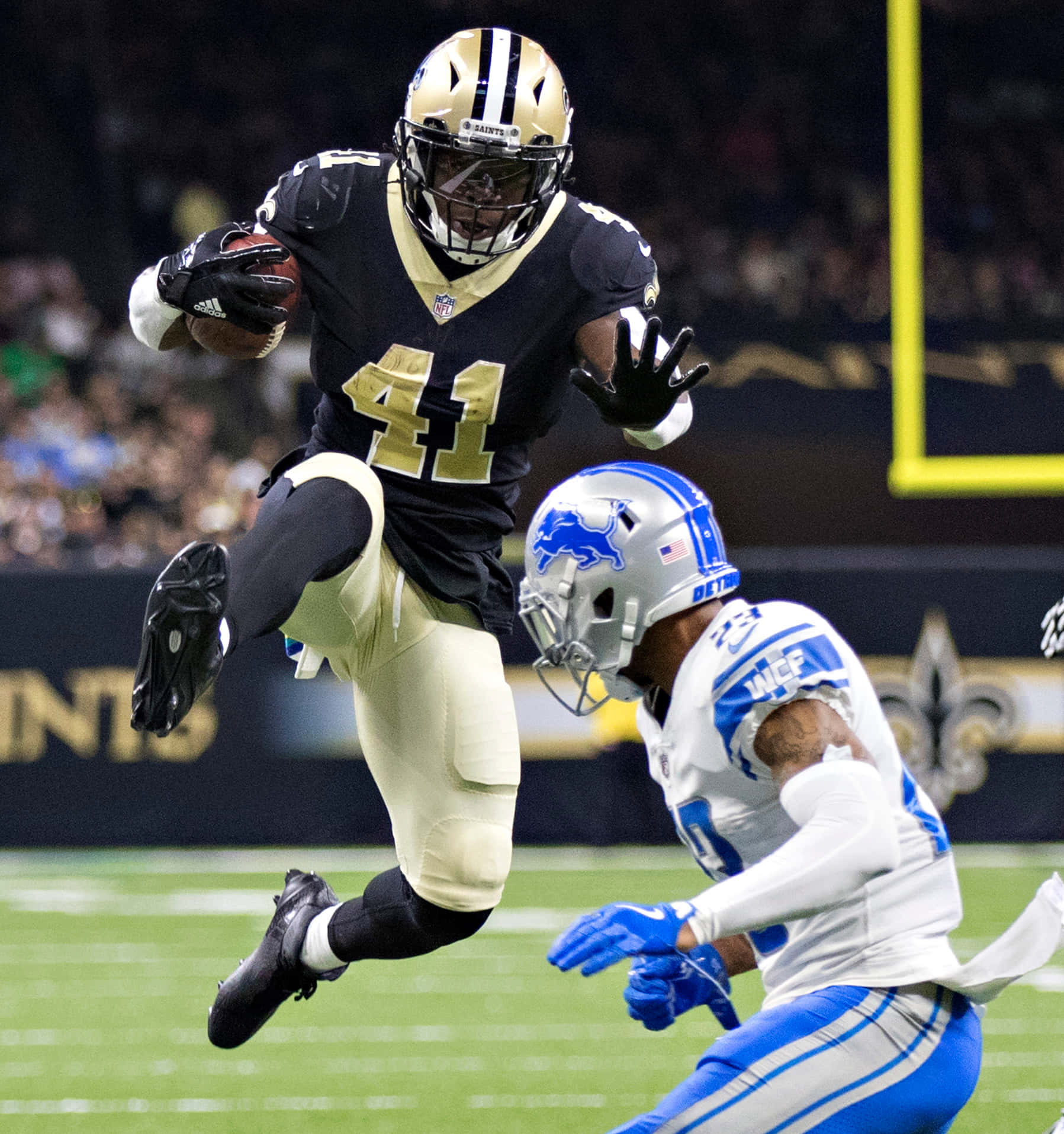 Alvin Kamara 桌布 壁紙 — 紐奧良聖徒隊球員在空中跳起抓住橄欖球，而底特律雄獅隊防守球員在燈光明亮的球場上飛身擒抱他。