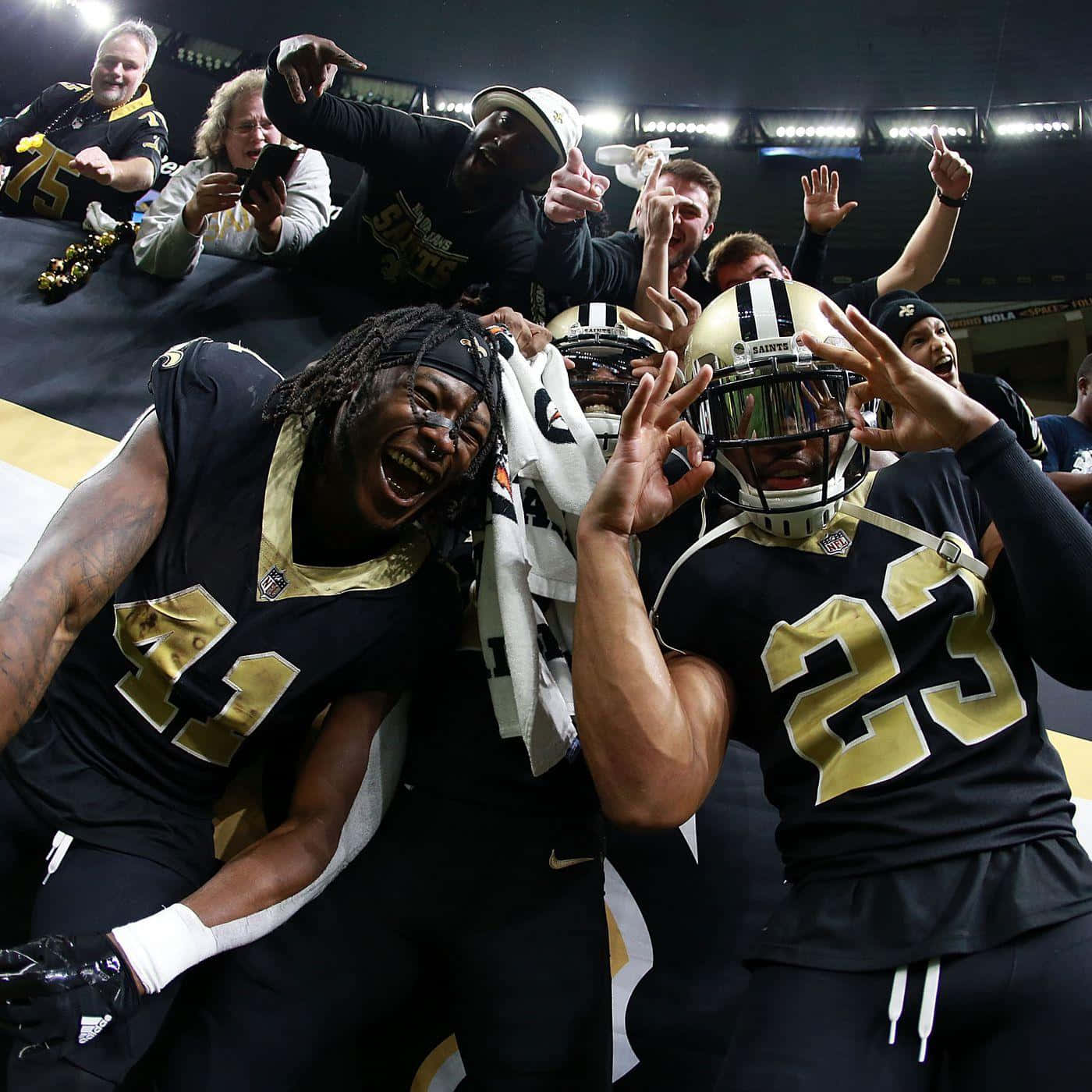 New Orleans Saints 桌布 壁紙 — 紐奧良聖徒隊的美式足球員在明亮的球場燈光下，在場上勝利慶祝，背景有球迷歡呼。