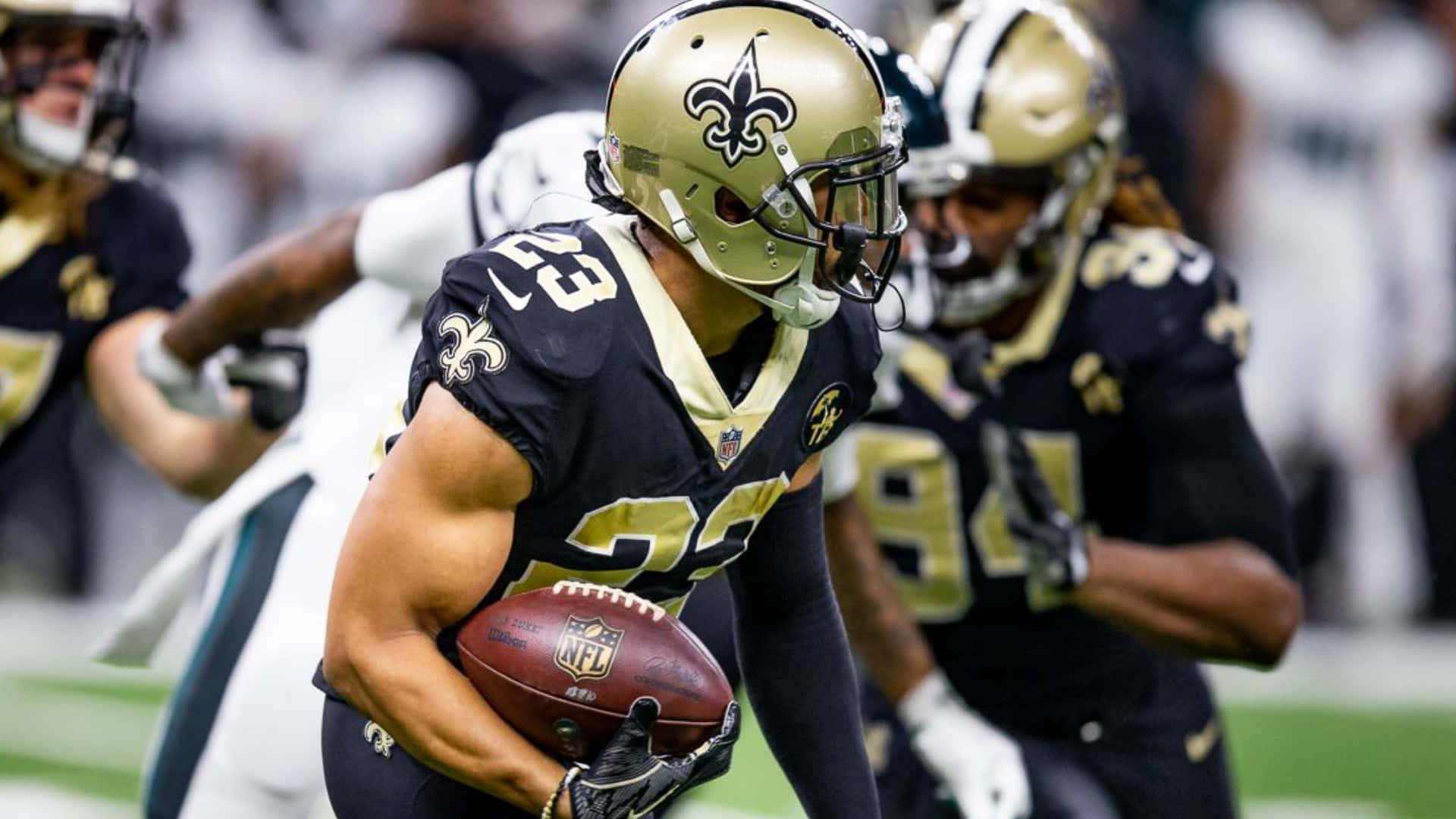 New Orleans Saints 桌布 壁紙 — 紐奧良聖徒隊的美式足球員身穿金色和黑色裝備，在體育場燈光下比賽中帶球奔跑。