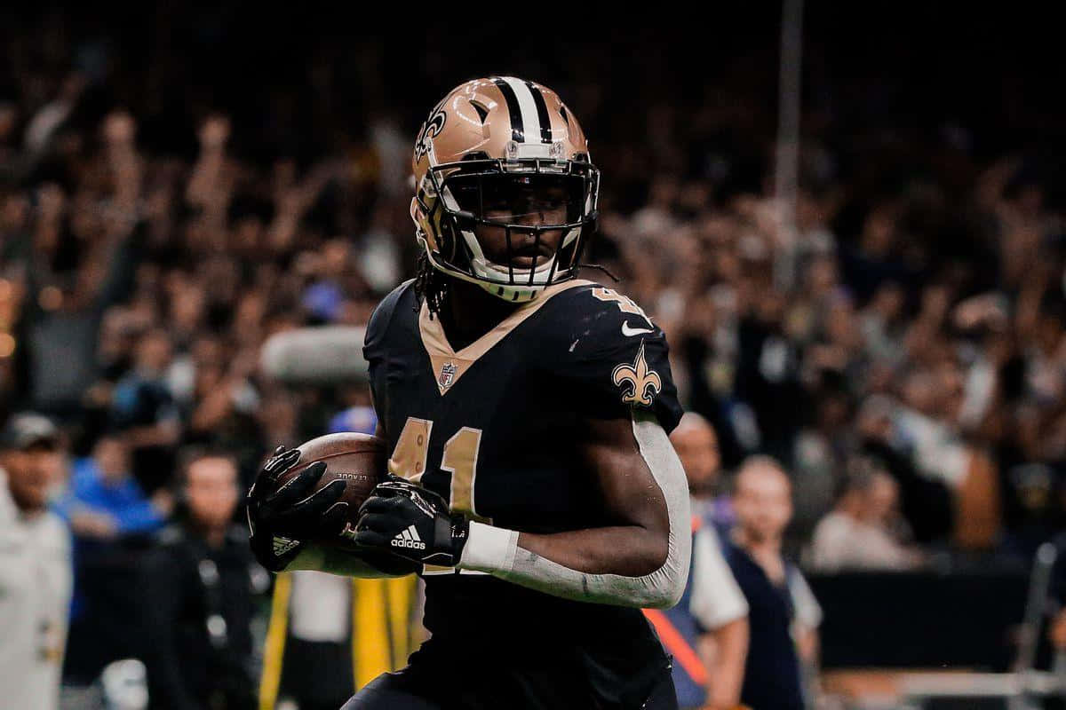 Alvin Kamara 桌布 壁紙 — 紐奧良聖徒隊的美式足球員身穿黑色和金色裝備，在球場燈光下帶球奔跑。