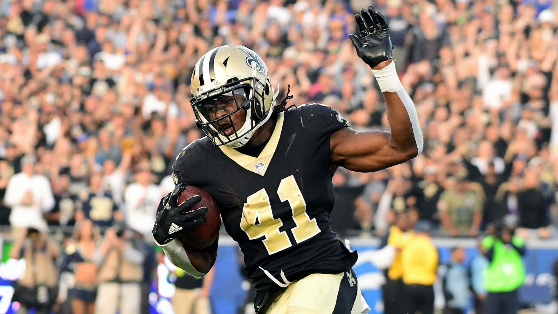 Alvin Kamara 桌布 壁紙 — 紐奧良聖徒隊的美式足球員身穿黑色和金色制服，帶著球奔跑，舉手在歡呼的人群前慶祝。