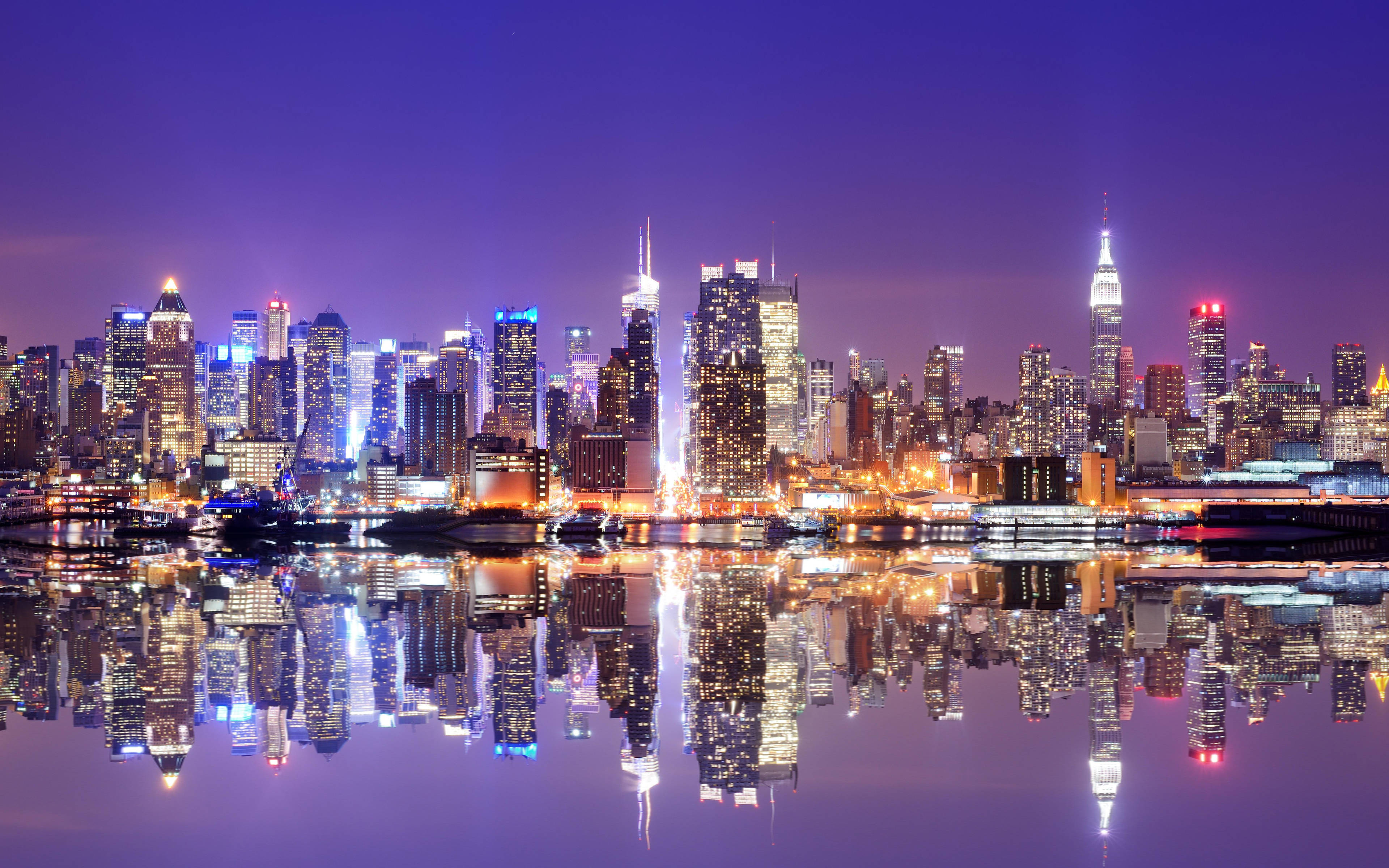 New York 桌布 壁紙 — 紐約令人目眩的夜景，摩天大樓在深紫色天空下閃耀，完美倒映在下方平靜的水面上。