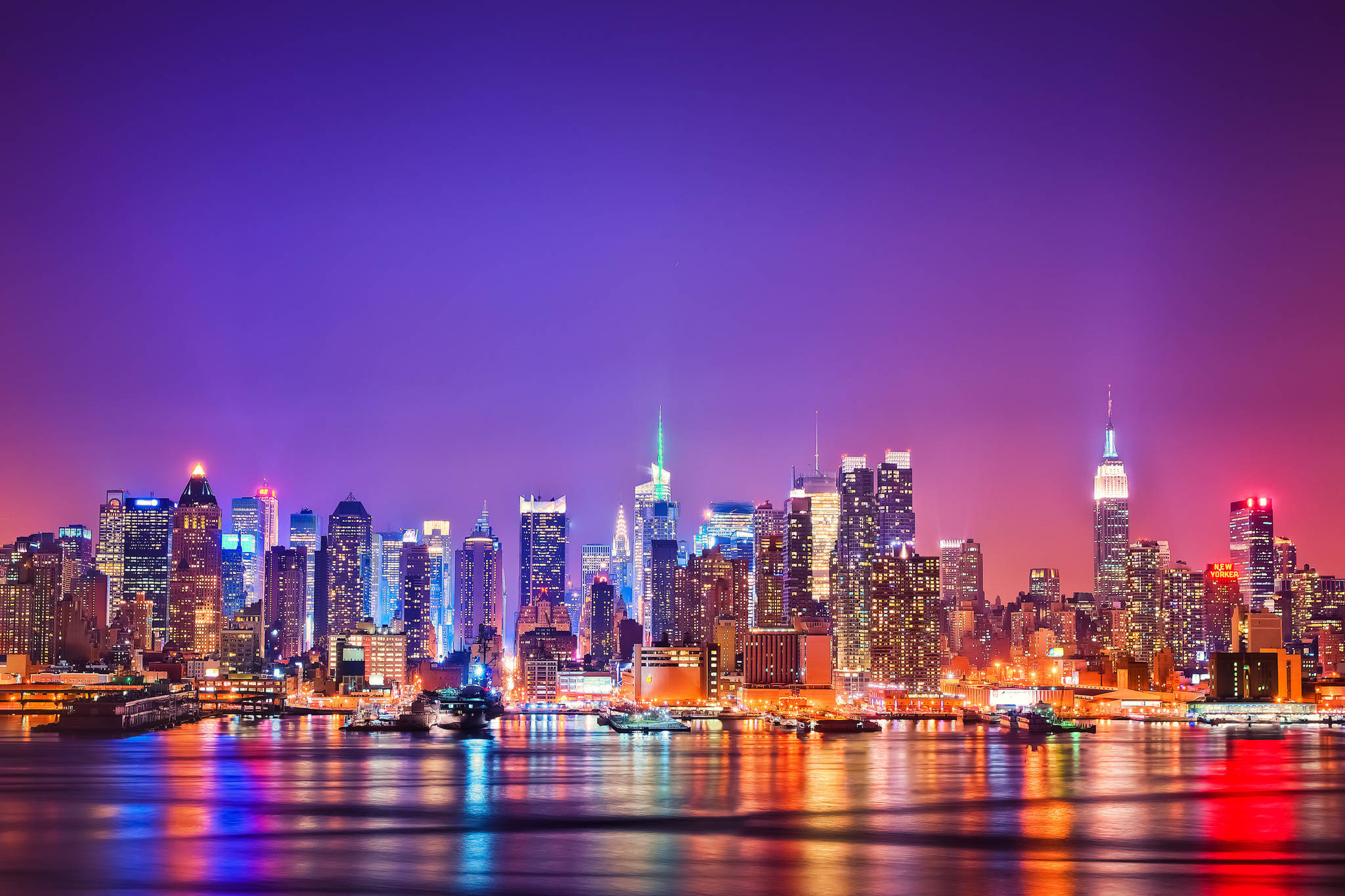 Nyc 桌布 壁紙 — 紐約令人目眩的夜景，摩天大樓在紫色、粉紅色和藍色燈光中閃耀，倒映在水面上。