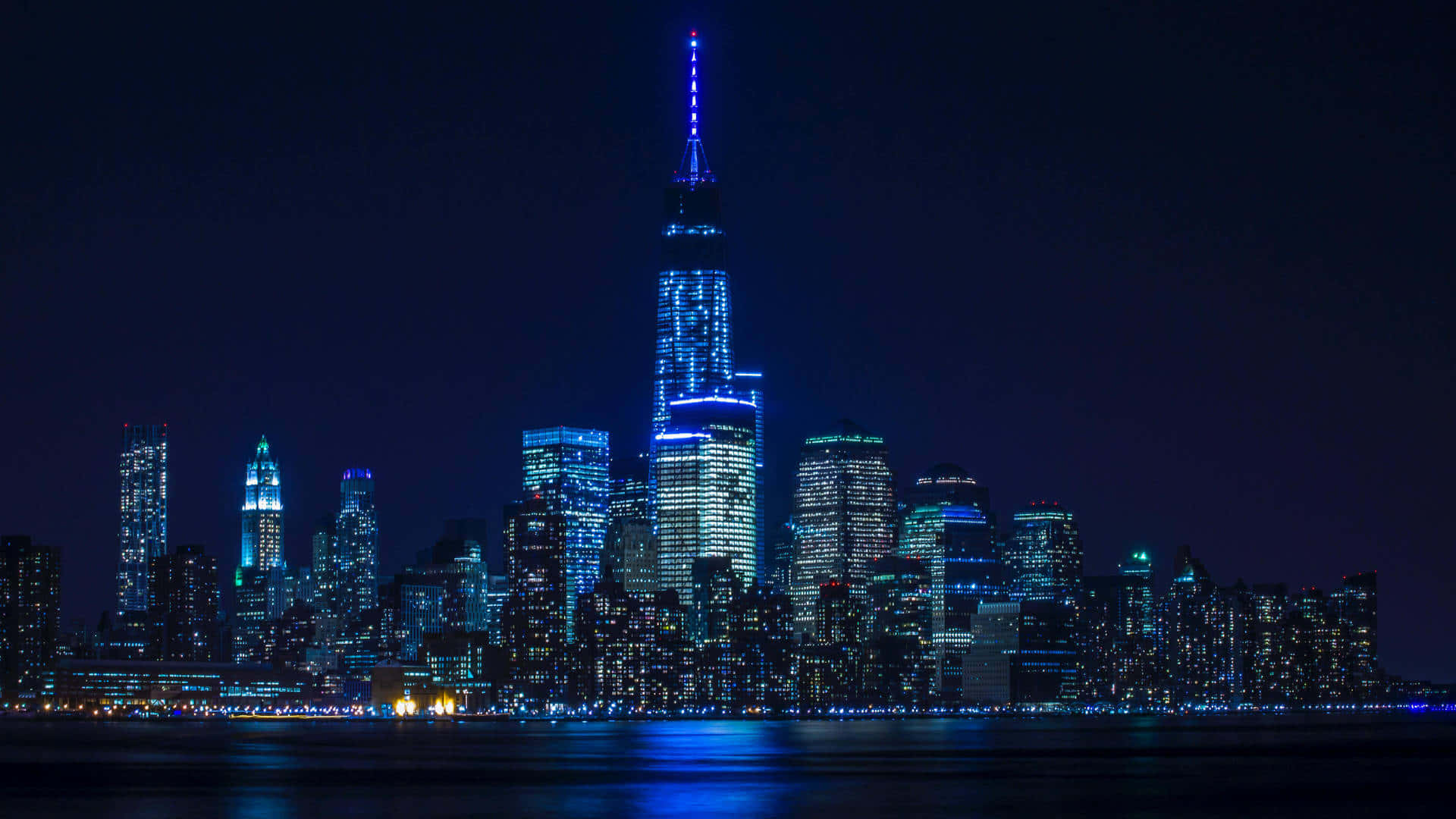 New York Skyline 桌布 壁紙 — 紐約令人目眩的夜景，摩天大樓在黑暗的天空中閃爍著藍色和白色的光芒，倒映在下方平靜的水面上。