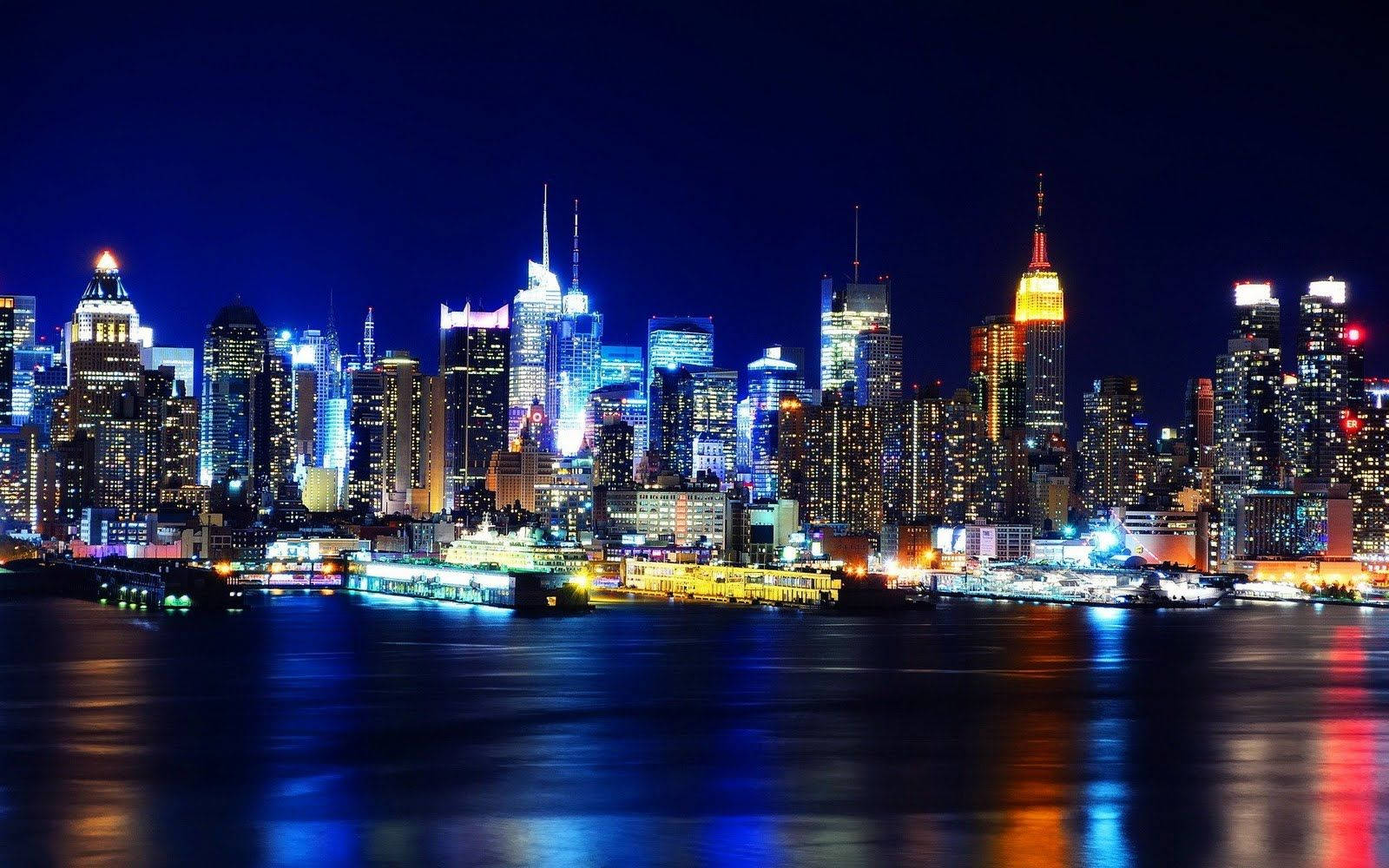 New York 桌布 壁紙 — 紐約令人目眩的夜景，摩天大樓在黑暗的天空中閃耀，水面上閃爍著充滿活力的倒影。