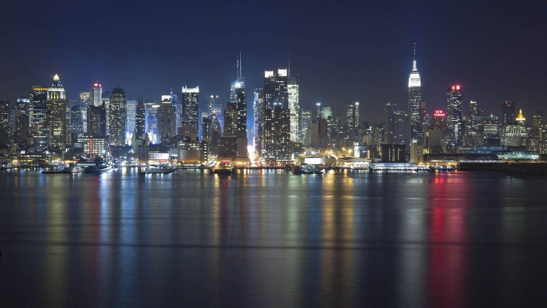 New York Skyline 桌布 壁紙 — 紐約令人目眩神迷的夜景，摩天大樓在漆黑的天空中閃耀，燈光倒映在下方平靜的水面上。