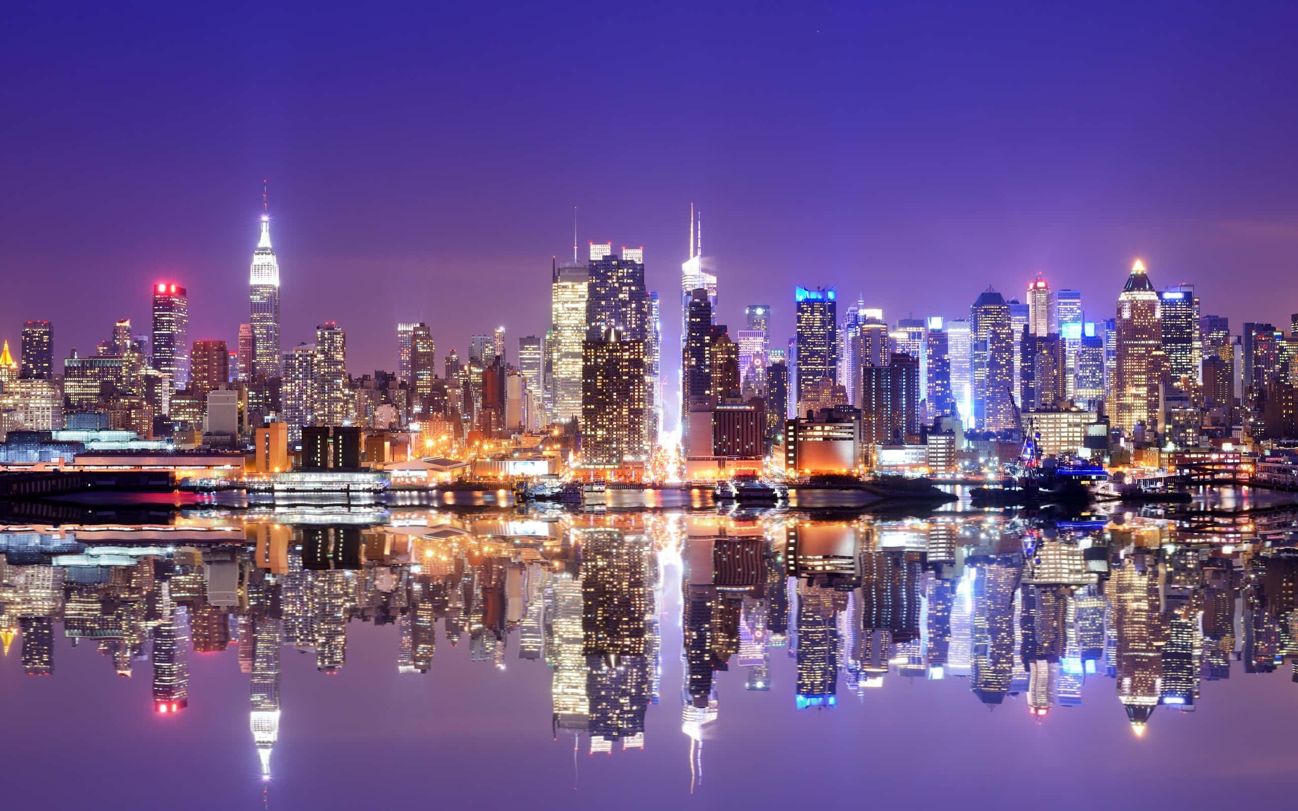 城市 桌布 壁紙 — 紐約令人眼花繚亂的夜景，摩天大樓在紫色天空下閃耀，完美倒映在下方平靜的水面上。