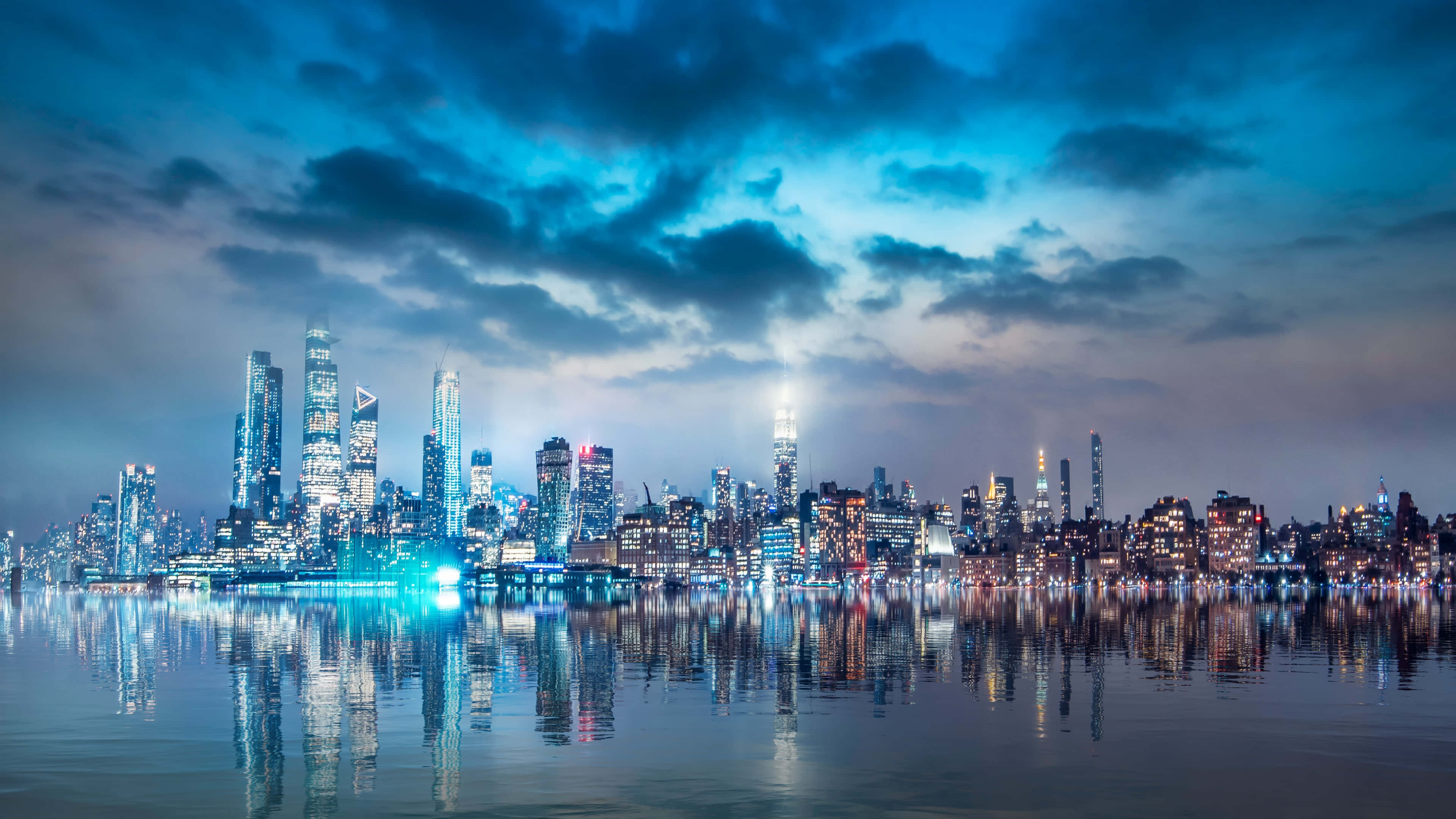 城市風景 桌布 壁紙 — 紐約令人驚嘆的夜景，摩天大樓在戲劇性的藍天下閃耀，倒映在平靜的水面上。
