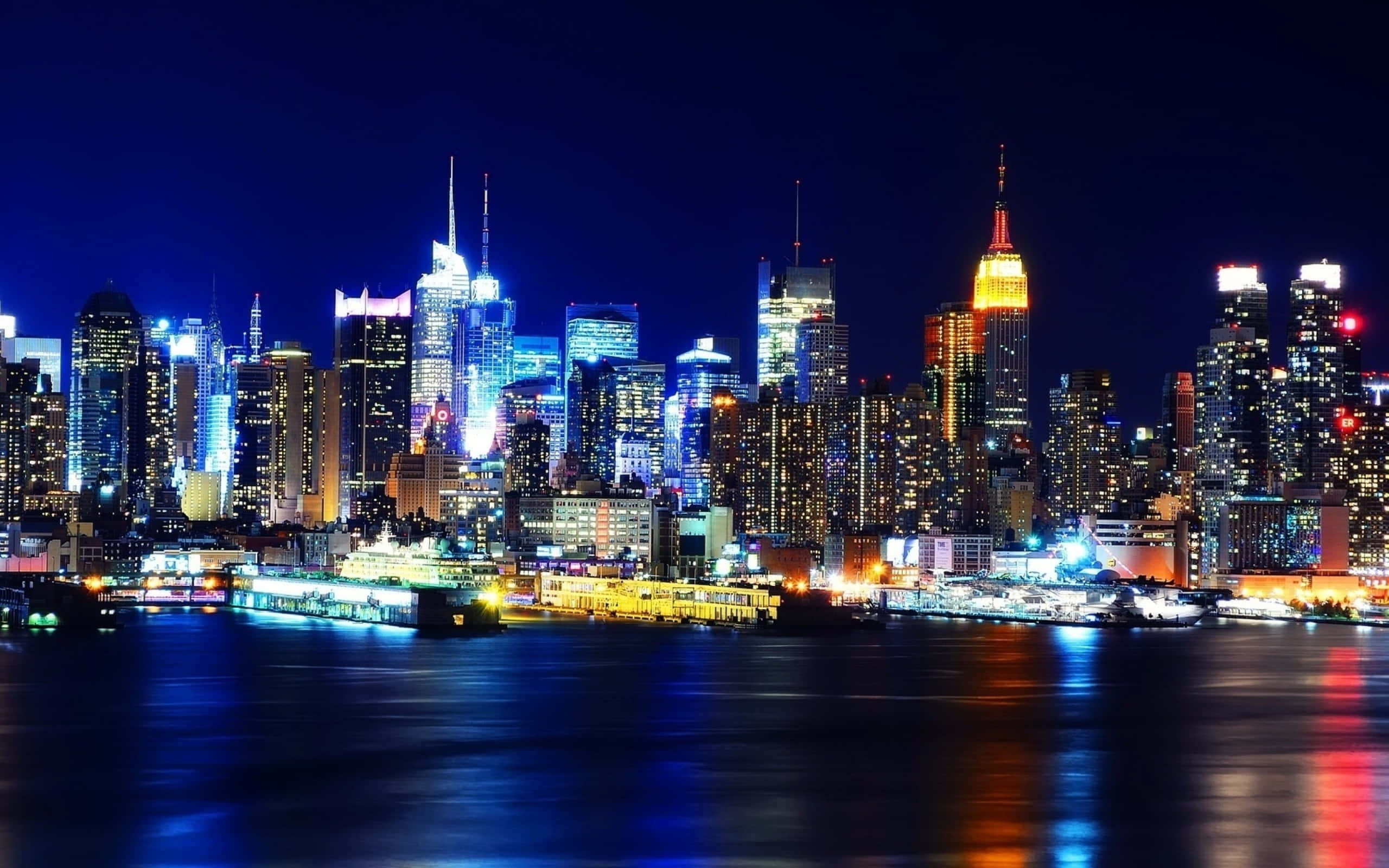 City Night 桌布 壁紙 — 紐約令人驚嘆的夜景，摩天大樓在黑暗的天空中閃耀，燈光倒映在下方平靜的水面上。