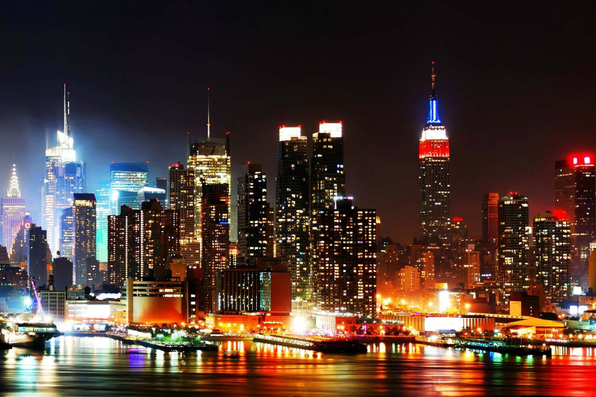 Nyc 桌布 壁紙 — 紐約令人驚嘆的夜景，摩天大樓在黑暗的天空中閃耀， vibrant 燈光在水面上反射。