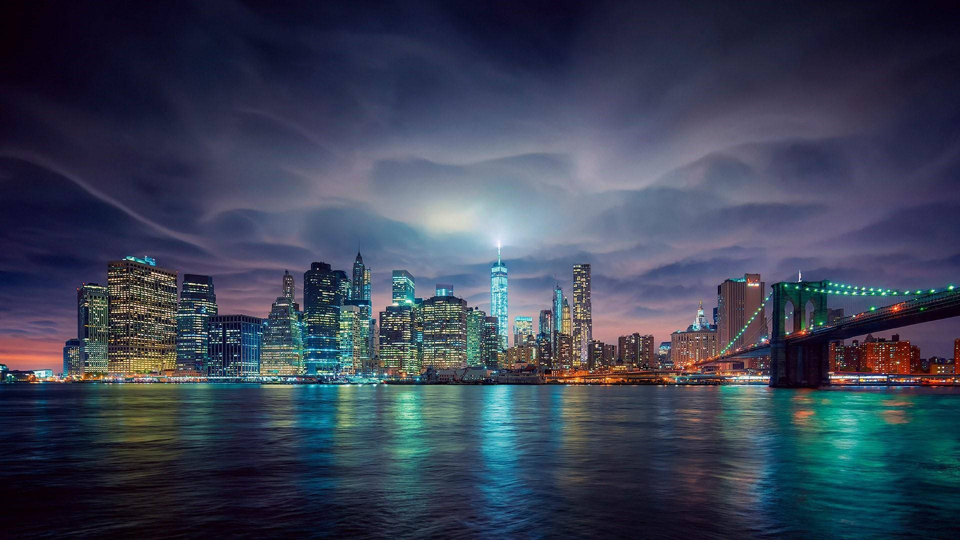 Skyscraper 桌布 壁紙 — 紐約充滿活力的夜景，閃爍的摩天大樓和布魯克林大橋倒映在平靜的水面上，天空戲劇性地多雲。