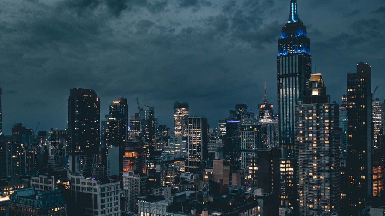 720p Empire State Building 桌布 壁紙 — 紐約夜晚的城市景觀，氣氛陰鬱，高聳的摩天大樓在黑暗多雲的天空下被溫暖的窗戶照亮。