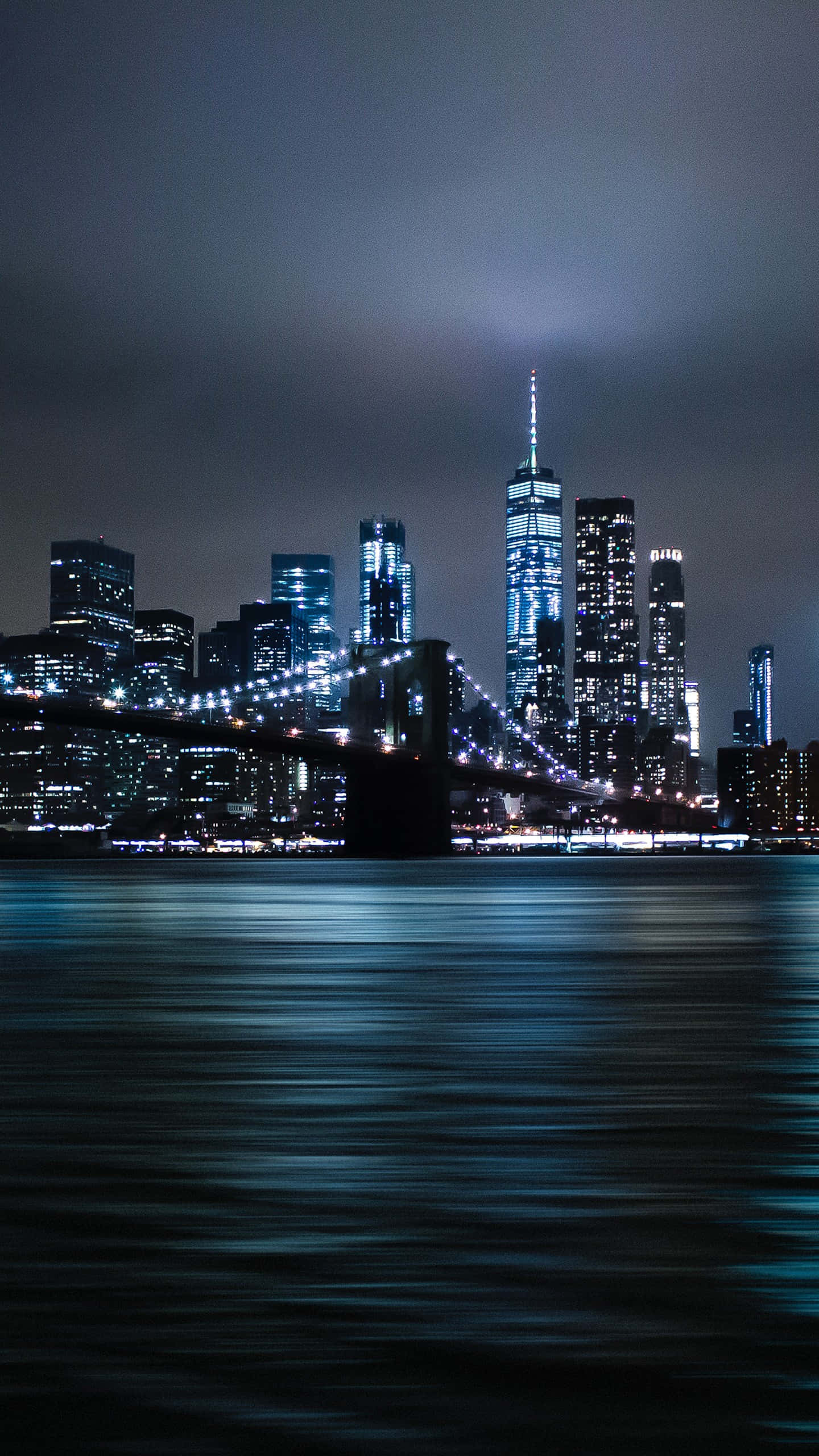 New York City Night iPhone 桌布 壁紙 — 紐約夜景，布魯克林大橋和世界貿易中心一號樓在黑暗天空下閃耀，倒映在平靜的水面上。