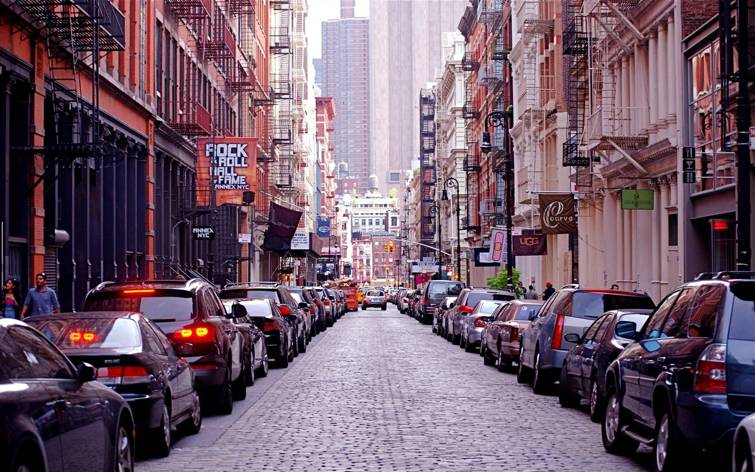 New York Street 桌布 壁紙 — 紐約市一條狹窄的鵝卵石街道，兩旁停滿汽車，磚砌建築設有防火梯。