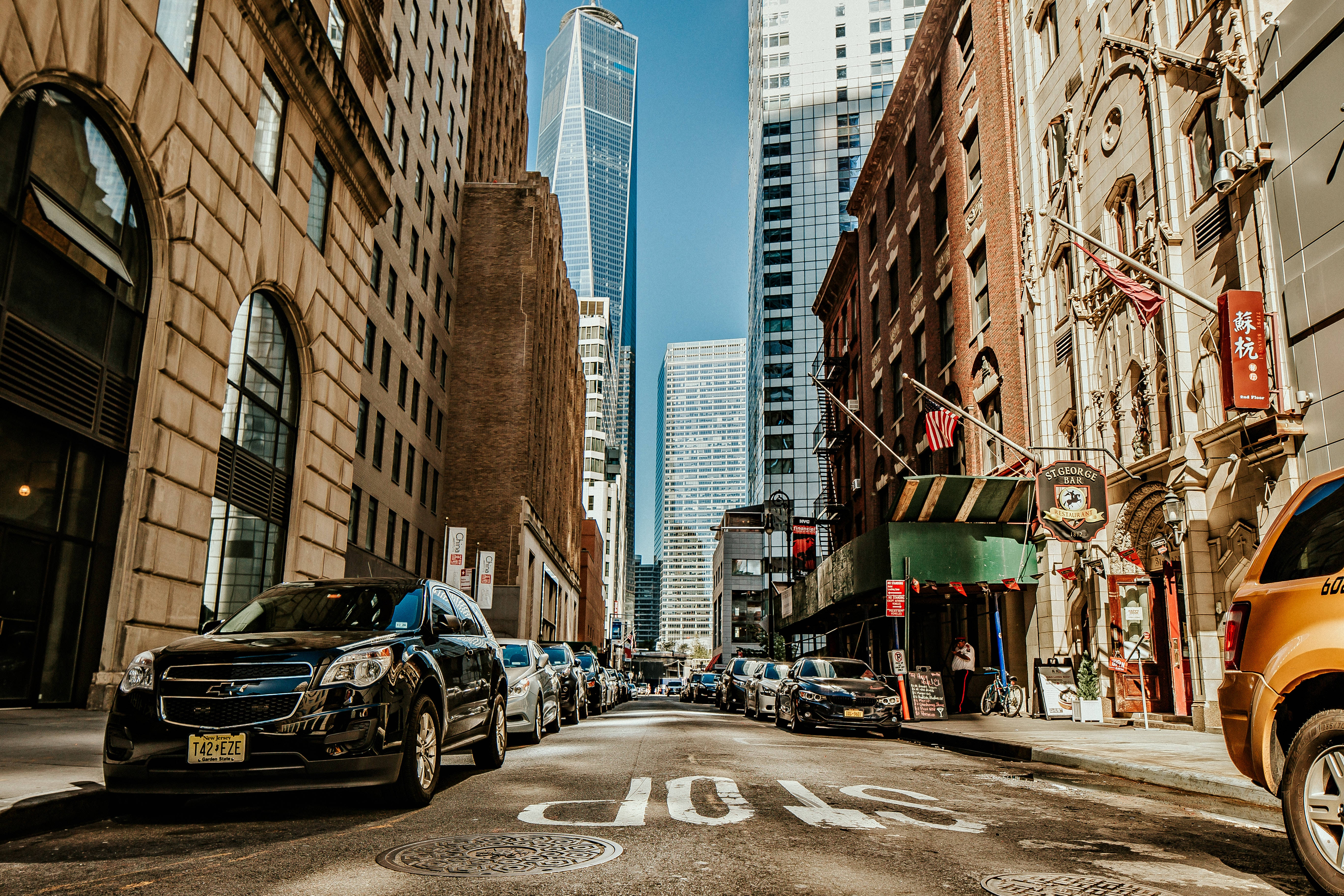 Buildings 桌布 壁紙 — 紐約市一條陽光普照的都市街道，兩旁是歷史悠久的磚造建築和高聳的玻璃摩天大樓，路邊停滿了汽車。