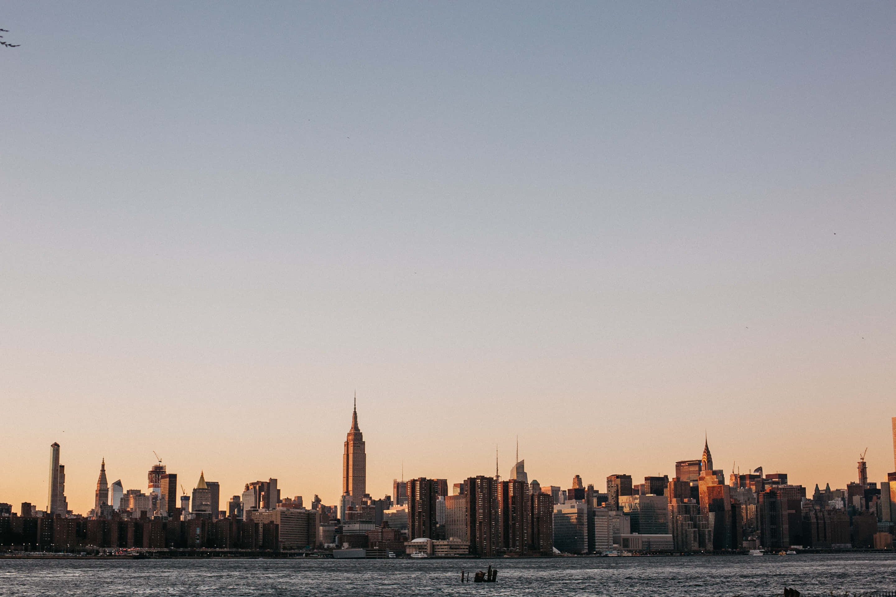 Nyc 桌布 壁紙 — 紐約市天際線在日落時分，金色光芒沐浴摩天大樓和下方平靜的水面。
