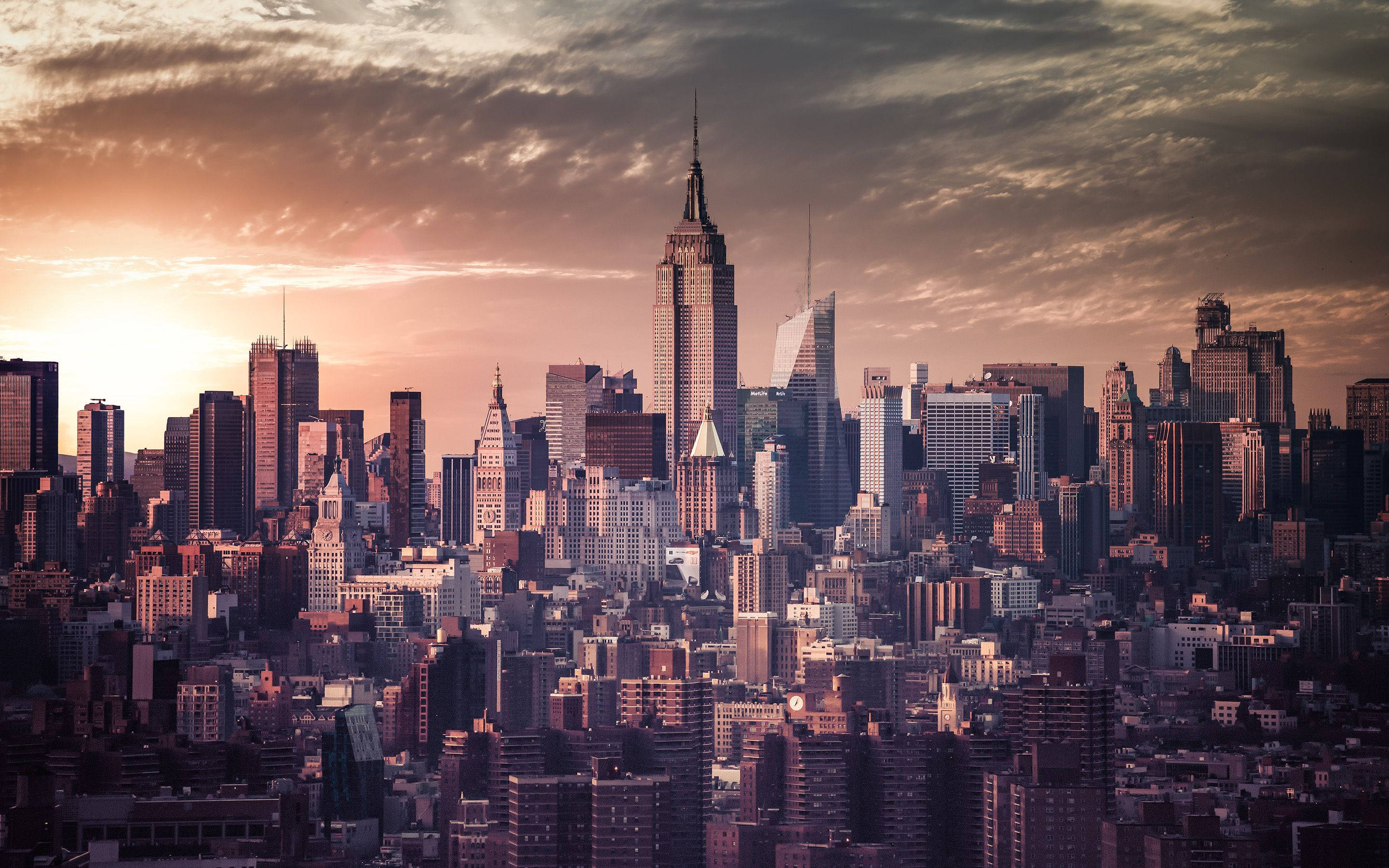 New York 桌布 壁紙 — 紐約市天際線在日落時的 panoram 景觀，金色光線照亮摩天大樓和戲劇性的雲層。