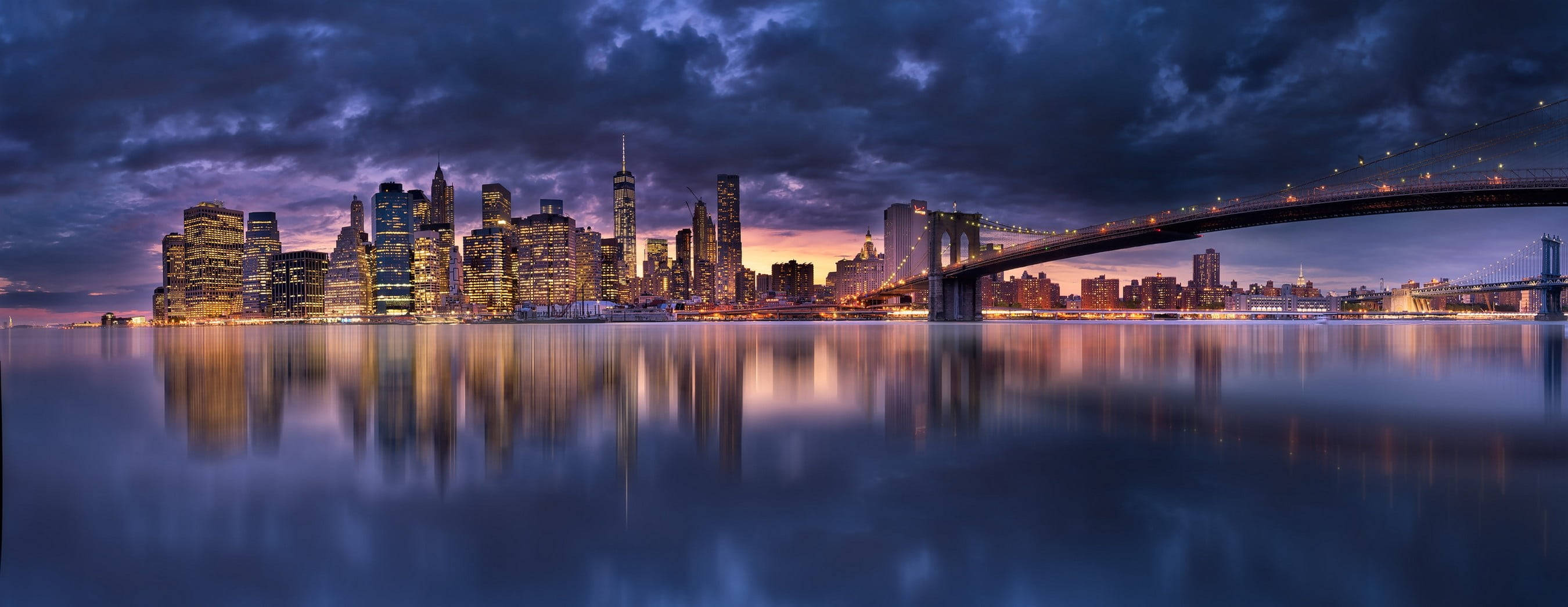 New York City Landscape 桌布 壁紙 — 紐約市天際線在黃昏時分的全景，燈火通明的摩天大樓和布魯克林大橋倒映在平靜的水面上，天空中有戲劇性的雲彩。