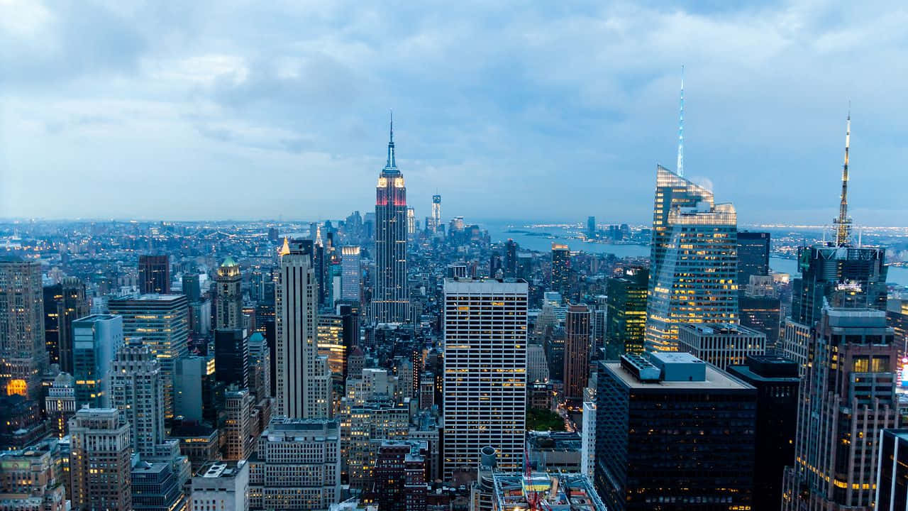 720p Empire State Building 桌布 壁紙 — 紐約市天際線在黃昏時分的全景視圖，高樓大廈燈火通明，天空多雲。