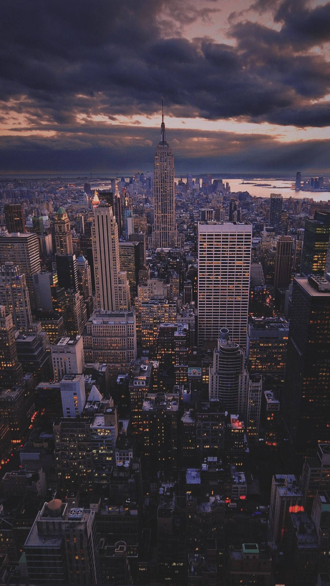 new york city iPhone x 桌布 壁紙 — 紐約市天際線在黃昏時的鳥瞰圖，帝國大廈在戲劇性的紫色雲層中亮起燈光。