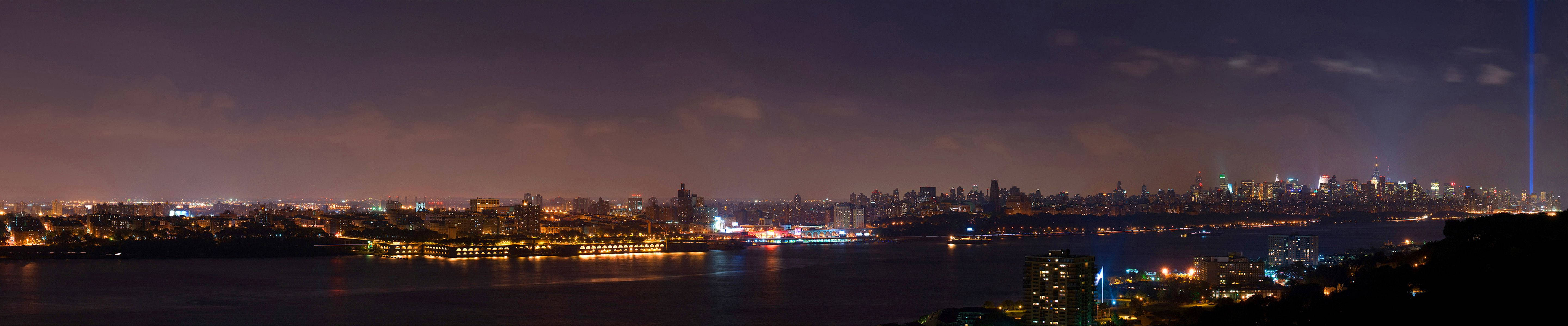 Triple 桌布 壁紙 — 紐約市天際線的全景夜景，發光的燈光在水面上反射，雙子星紀念光束刺穿天空。
