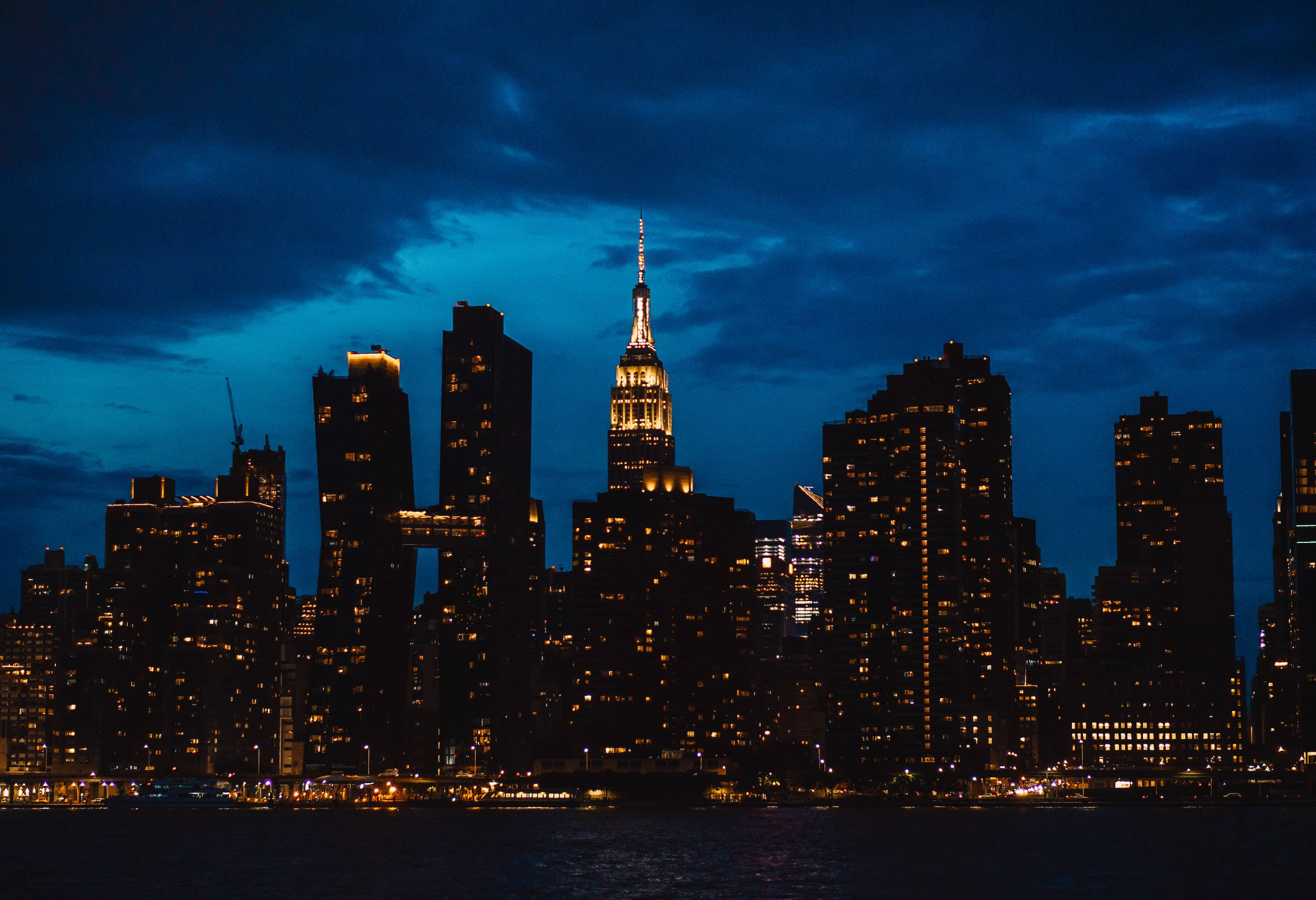Night Aesthetic 桌布 壁紙 — 紐約市天際線的壯麗夜景，摩天大樓燈火通明，在深藍色天空下倒映在水面上。