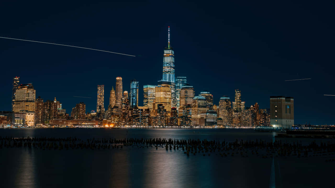 720p Ny City 桌布 壁紙 — 紐約市天際線的壯麗夜景，燈火通明的摩天大樓在平靜的水面上倒映，黑暗的天空中有飛機尾跡。