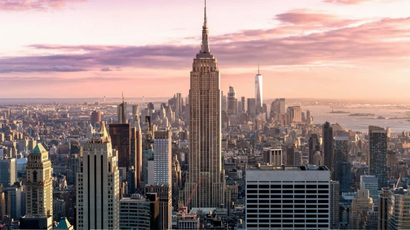 1366x768 Empire State Building 桌布 壁紙 — 紐約市天際線的壯麗日落景觀，帝國大廈聳立在密集的城市建築和遠處的水面上。