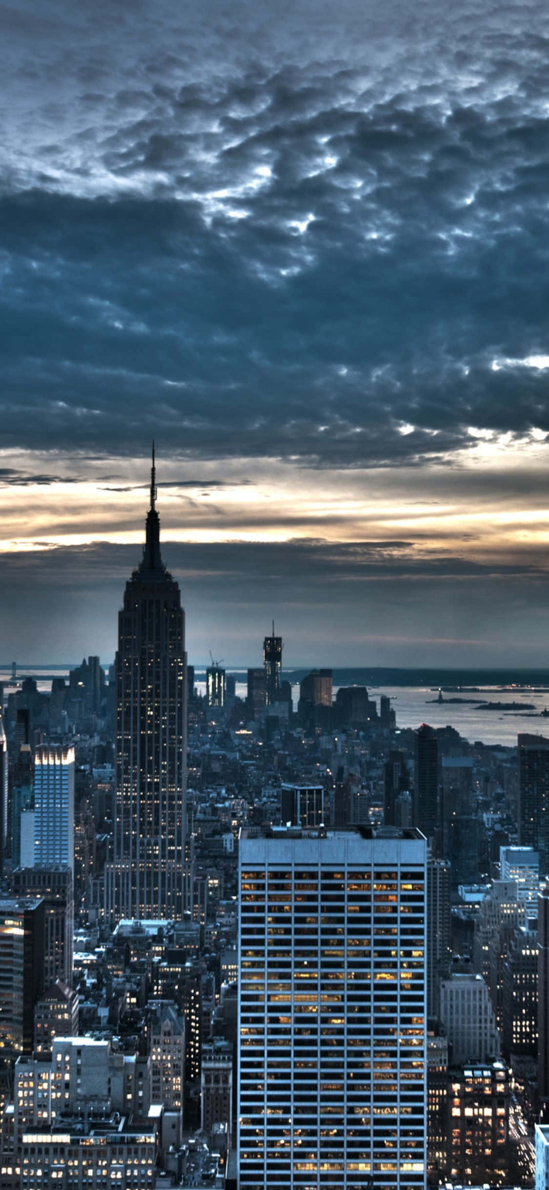 iPhone Xs Ny City 桌布 壁紙 — 紐約市天際線的戲劇性暮色景觀，帝國大廈在陰鬱的雲層和遠處的水面上亮起燈光。