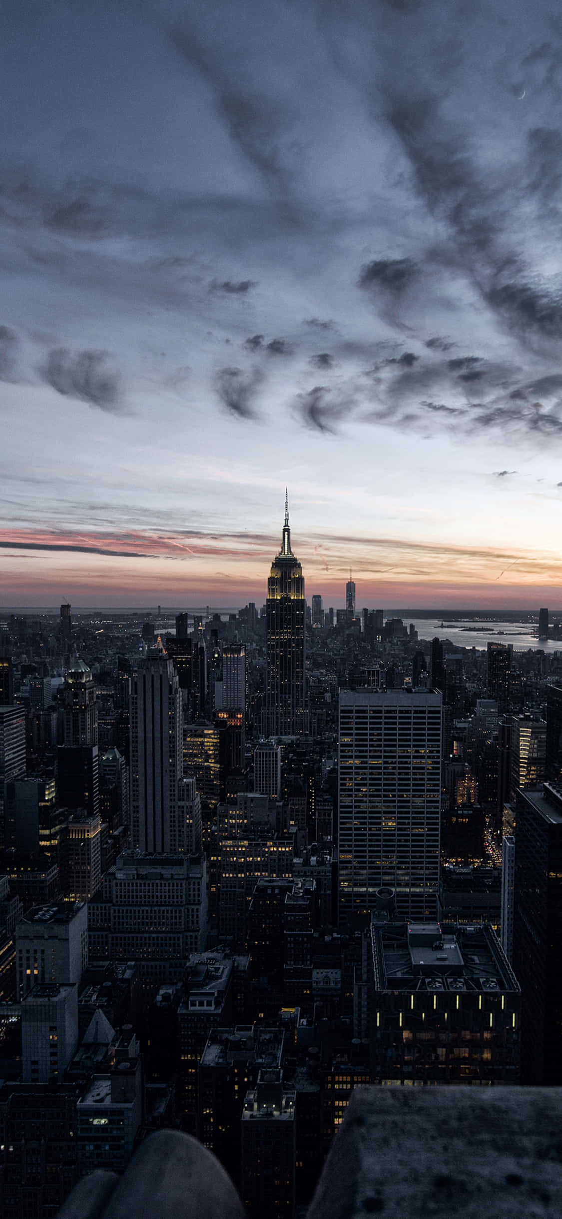 iPhone Xs New York 桌布 壁紙 — 紐約市天際線的戲劇性暮色景觀，帝國大廈在陰鬱的雲層和夕陽餘暉中閃耀。