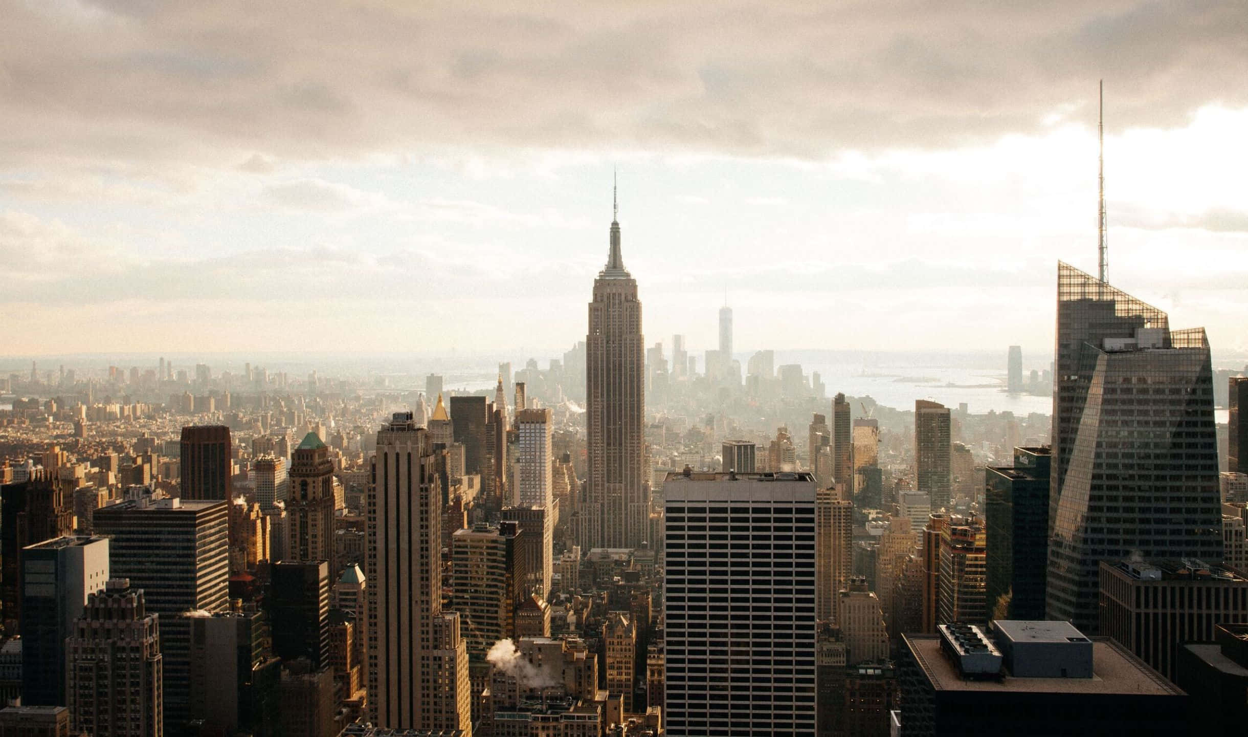 2440x1440 Empire State Building 桌布 壁紙 — 紐約市天際線的日出全景，帝國大廈在密集的摩天大樓之上聳立，沐浴在柔和的金色光芒中。